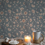 Cozy and intimate corner featuring Eternal Spring Wallpaper with a pattern of beige floral blooms linked by dark blue vines on a muted teal background. A candle lit on a wooden tray beside a ceramic mug and white crystals on a plate adds a warm, inviting ambiance.