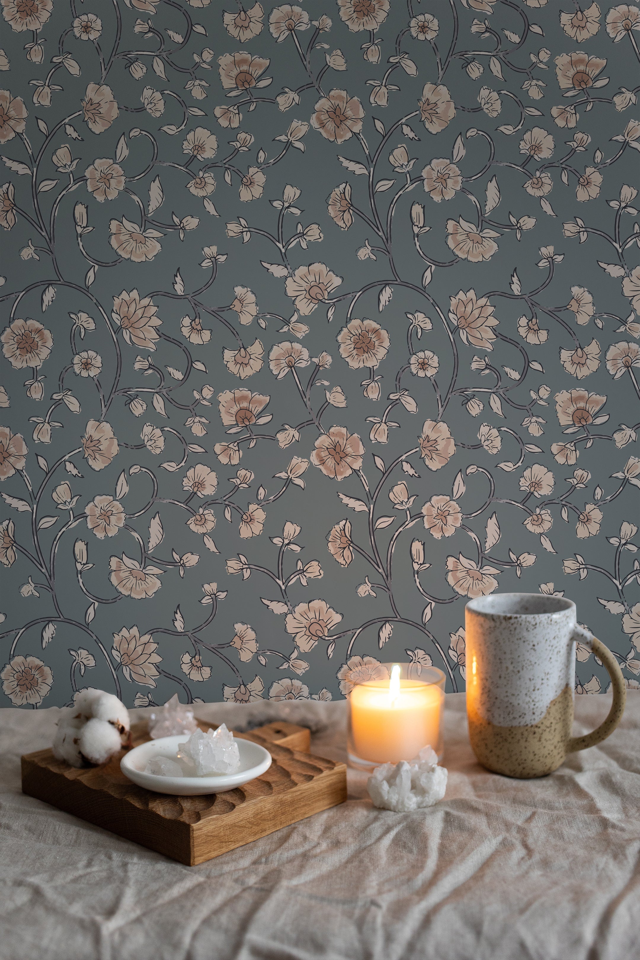 Cozy and intimate corner featuring Eternal Spring Wallpaper with a pattern of beige floral blooms linked by dark blue vines on a muted teal background. A candle lit on a wooden tray beside a ceramic mug and white crystals on a plate adds a warm, inviting ambiance.