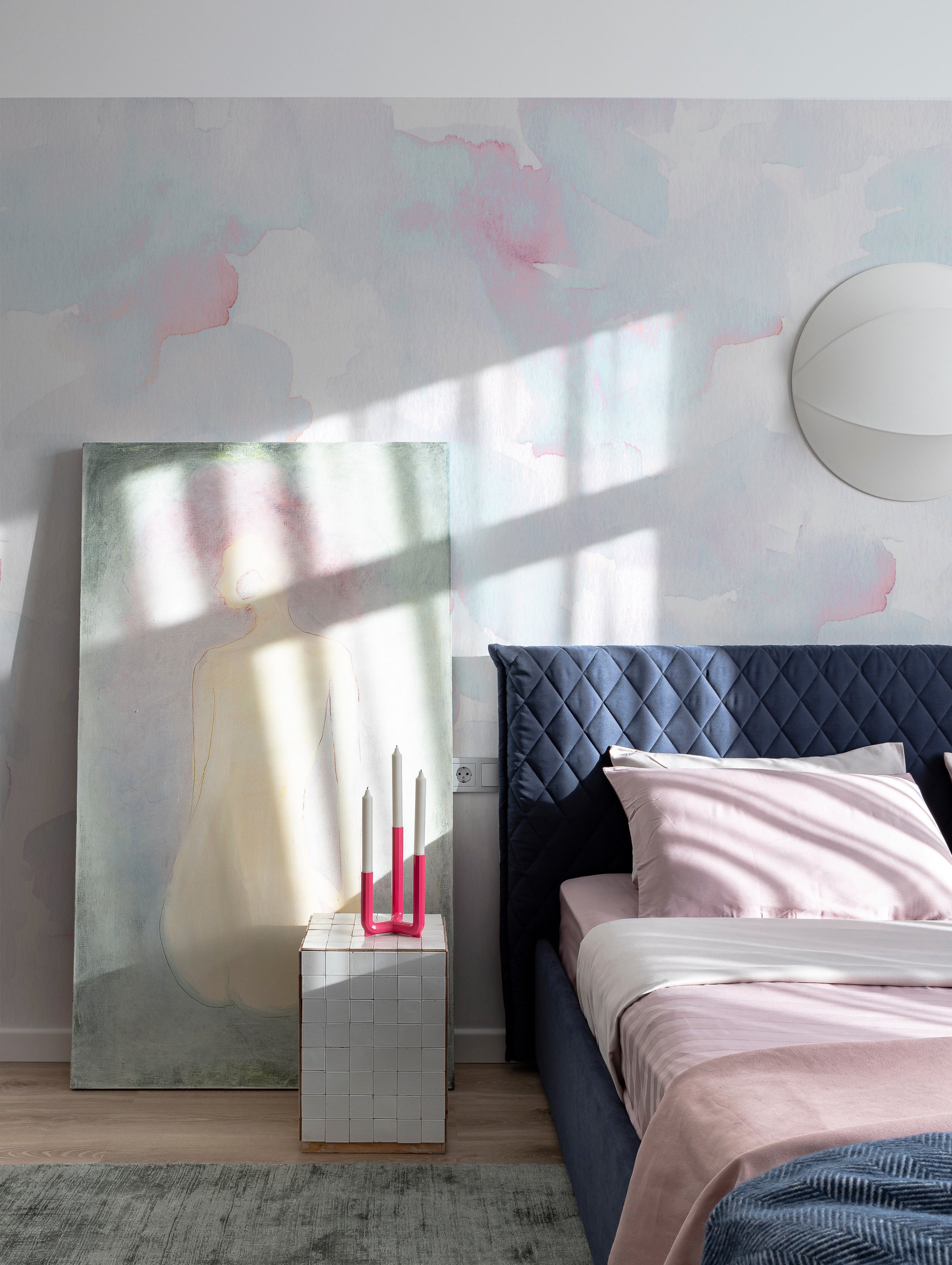Modern bedroom with the Hand Painted Mural - Cotton Candy as a focal wall, displaying ethereal watercolor patches in pink and blue. The room is styled with a navy upholstered bed, pink bedding, and a contemporary mirrored side table with pink candles, offering a chic and serene sleeping space.