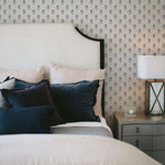 Elegant bedroom setup showing a large bed with a curved headboa0rd against a wall adorned with Tiny Hydrangea wallpaper. The bed is accessorized with dark blue and beige pillows, a side table with a lamp and a decorative box, creating a serene and sophisticated ambiance.