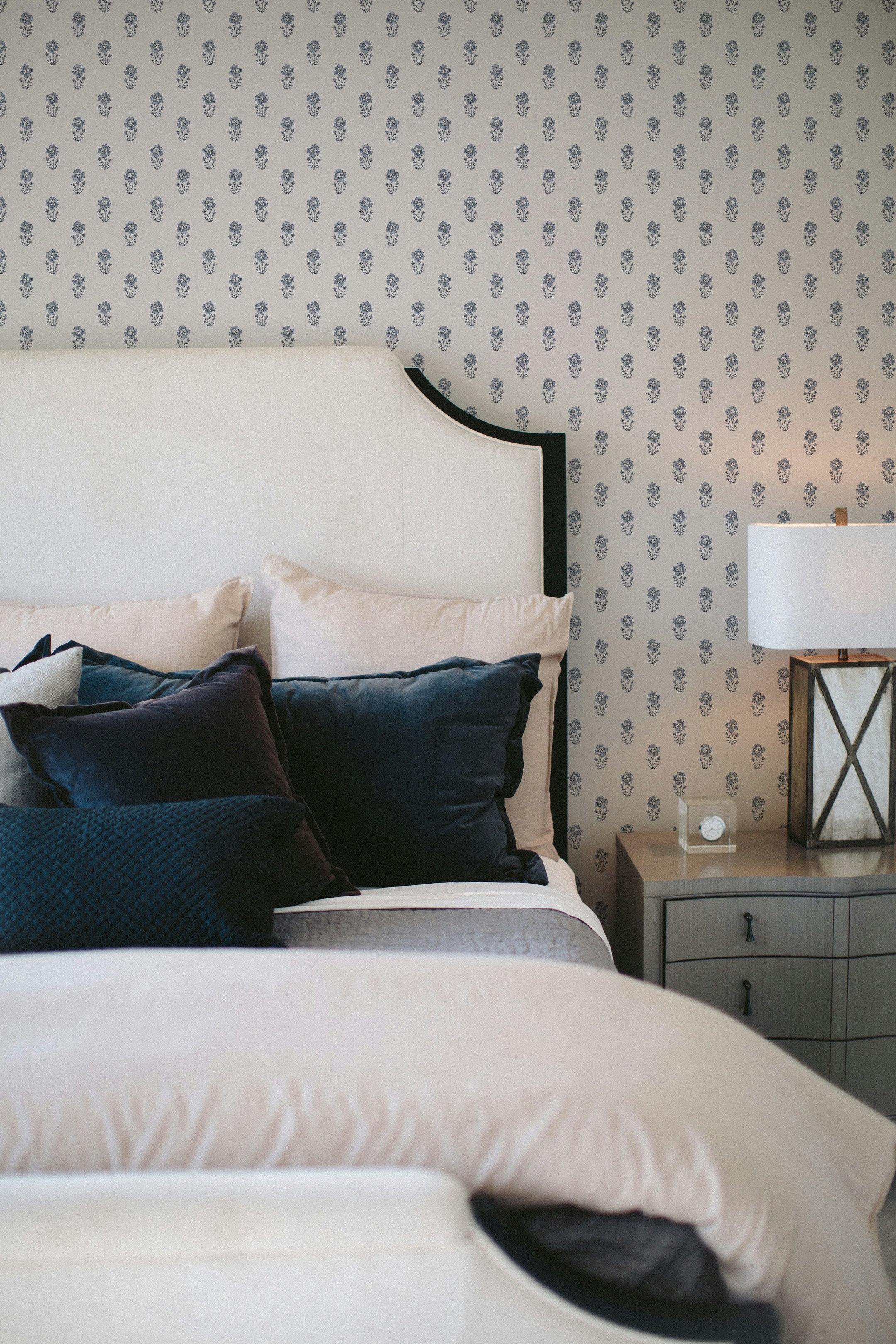 Elegant bedroom setup showing a large bed with a curved headboa0rd against a wall adorned with Tiny Hydrangea wallpaper. The bed is accessorized with dark blue and beige pillows, a side table with a lamp and a decorative box, creating a serene and sophisticated ambiance.