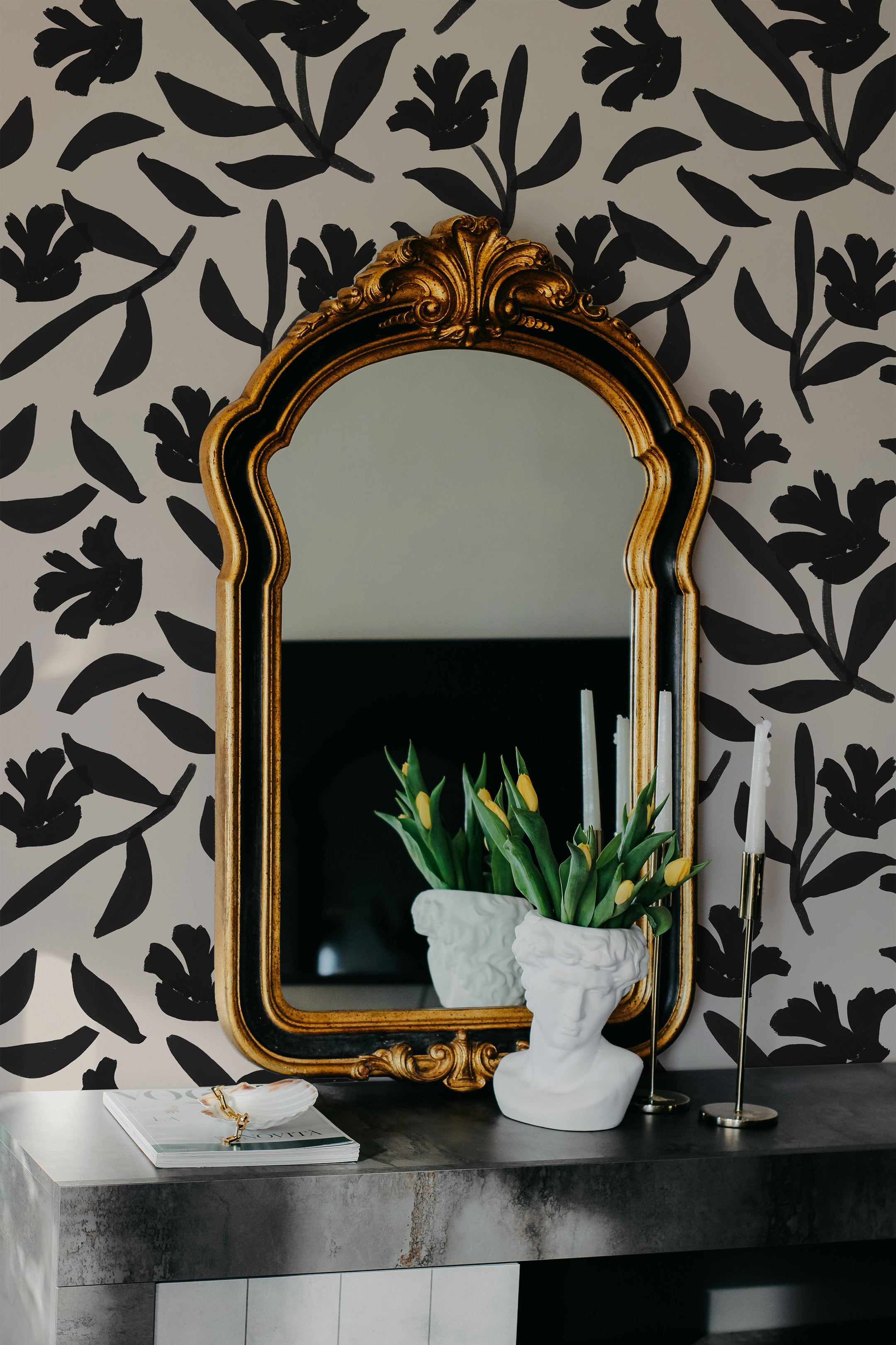 Chic room detail showcasing a black floral wallpaper with a large golden mirror above a modern grey console table. The table is decorated with white bust sculptures, yellow tulips in a white vase, and tall white candles, reflecting a sophisticated interior style