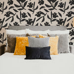 Elegant bedroom featuring a wall covered with a black floral pattern on a beige background. The bed is adorned with pillows in shades of yellow, grey, and a black and white pattern, creating a stylish and inviting atmosphere.