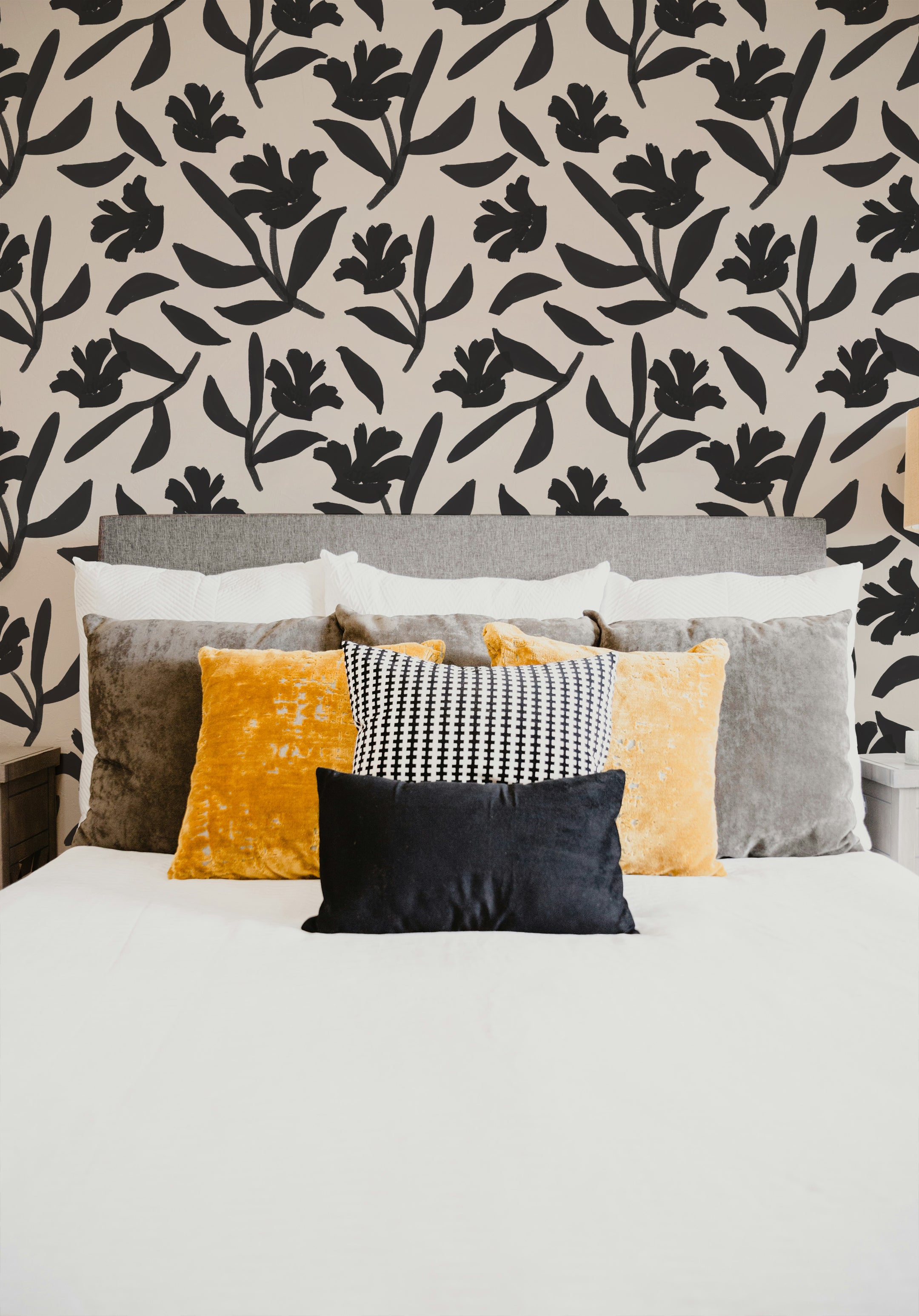 Elegant bedroom featuring a wall covered with a black floral pattern on a beige background. The bed is adorned with pillows in shades of yellow, grey, and a black and white pattern, creating a stylish and inviting atmosphere.