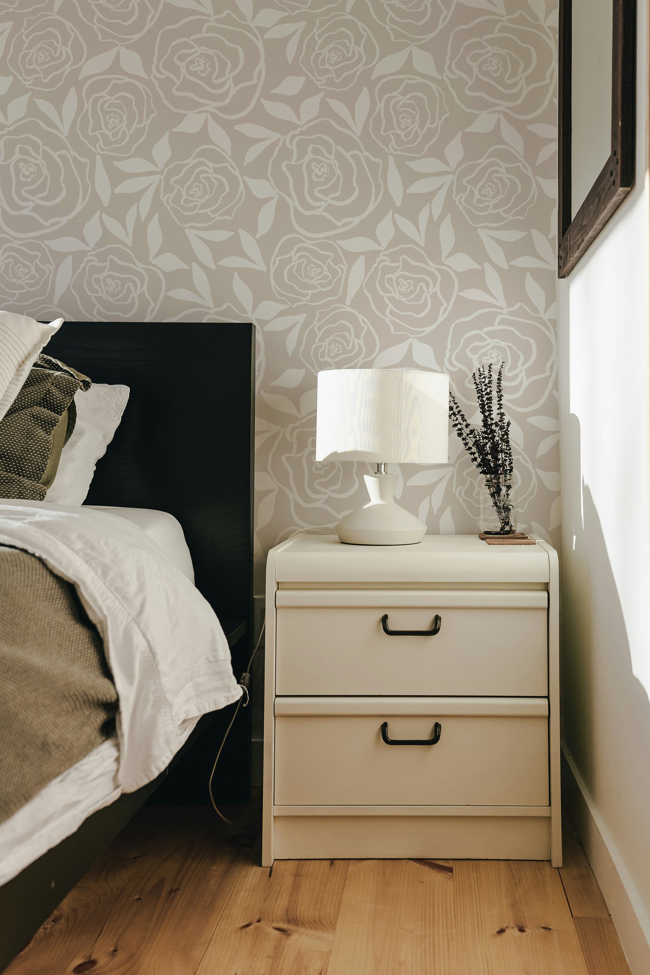 Charming bedroom scene with a rose-patterned wallpaper providing a serene backdrop. A bedside table with a white lamp and a decorative vase holding dried lavender next to a bed with linen bedding, creating a tranquil and elegant setting.
