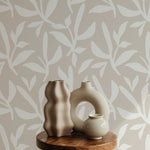 Detail view of a serene room corner with a wall covered in beige and white leaf patterned wallpaper. Artistic ceramic vases on a round wooden side table add a touch of elegance and simplicity to the minimalist decor.