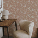 A cozy corner of a room featuring a plush beige chair and a wooden table with a lamp, set against a backdrop of soft pink wallpaper with white floral patterns, conveying a warm and inviting atmosphere.