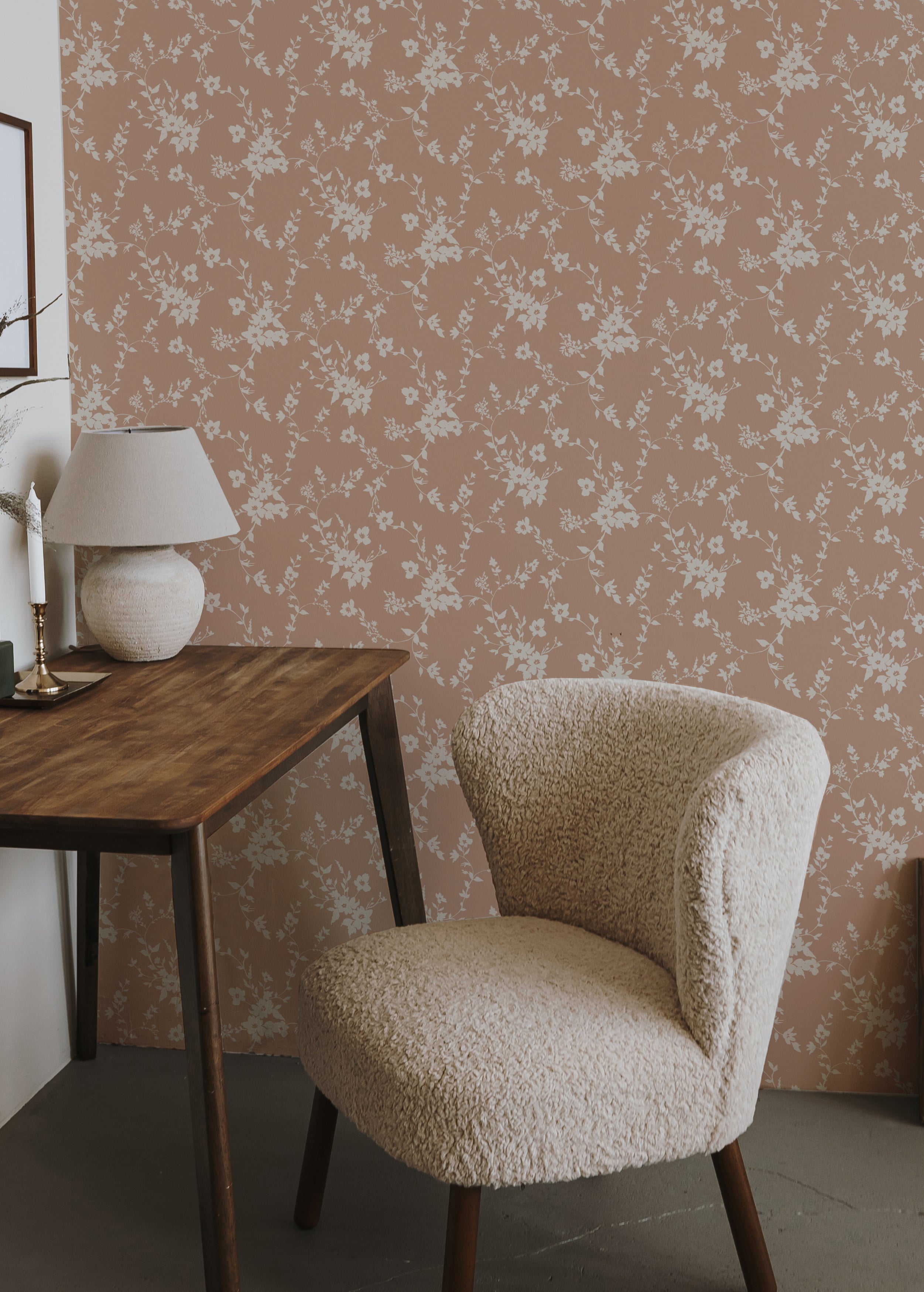 A cozy corner of a room featuring a plush beige chair and a wooden table with a lamp, set against a backdrop of soft pink wallpaper with white floral patterns, conveying a warm and inviting atmosphere.
