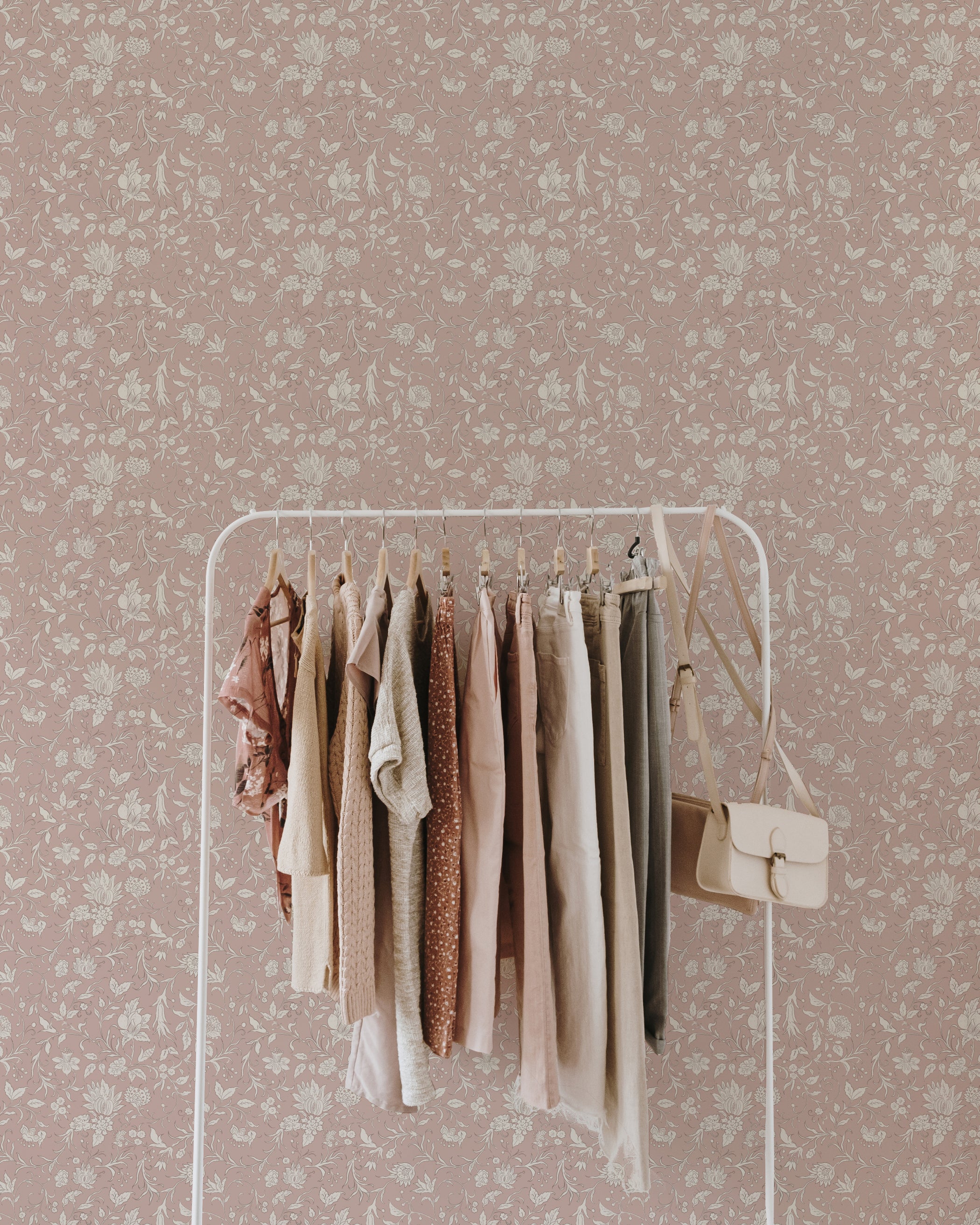 A white clothing rack filled with pastel-colored garments stands against a wall adorned with Noble Florals Wallpaper. The wallpaper features a delicate floral pattern in muted shades of mauve and cream, adding a vintage charm to the space.