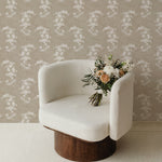 A cozy corner showcasing a modern, white textured armchair placed in front of a wall adorned with classic floral chintz wallpaper in white on a neutral background. A bouquet of fresh flowers adds a touch of color and elegance to the scene.