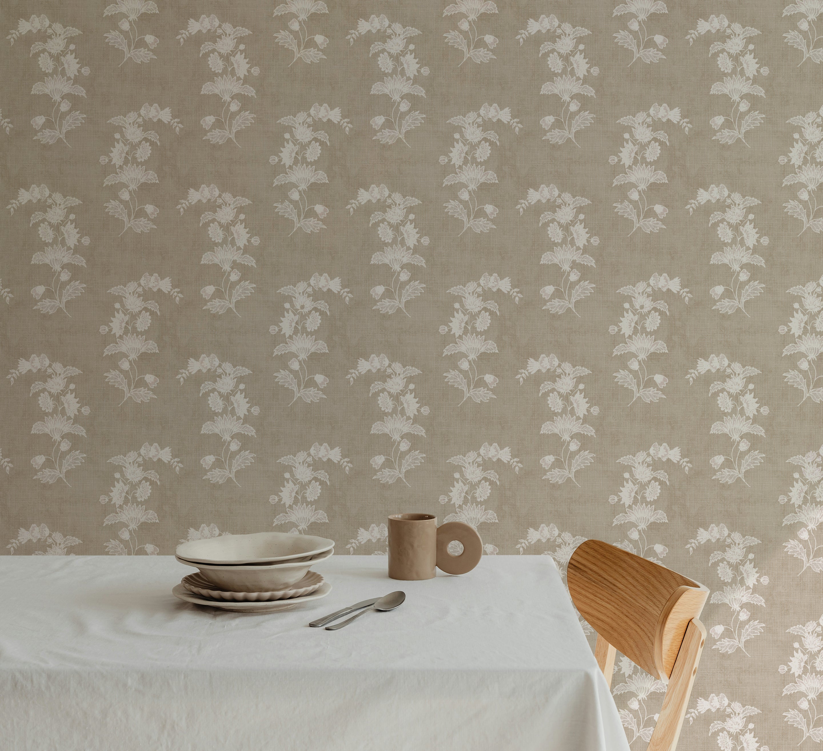 A dining setup featuring a simple white tablecloth and minimalist tableware on a rustic wooden table, set against a backdrop of a wall covered in a floral chintz wallpaper with white blossoms on a linen background, creating a serene dining atmosphere