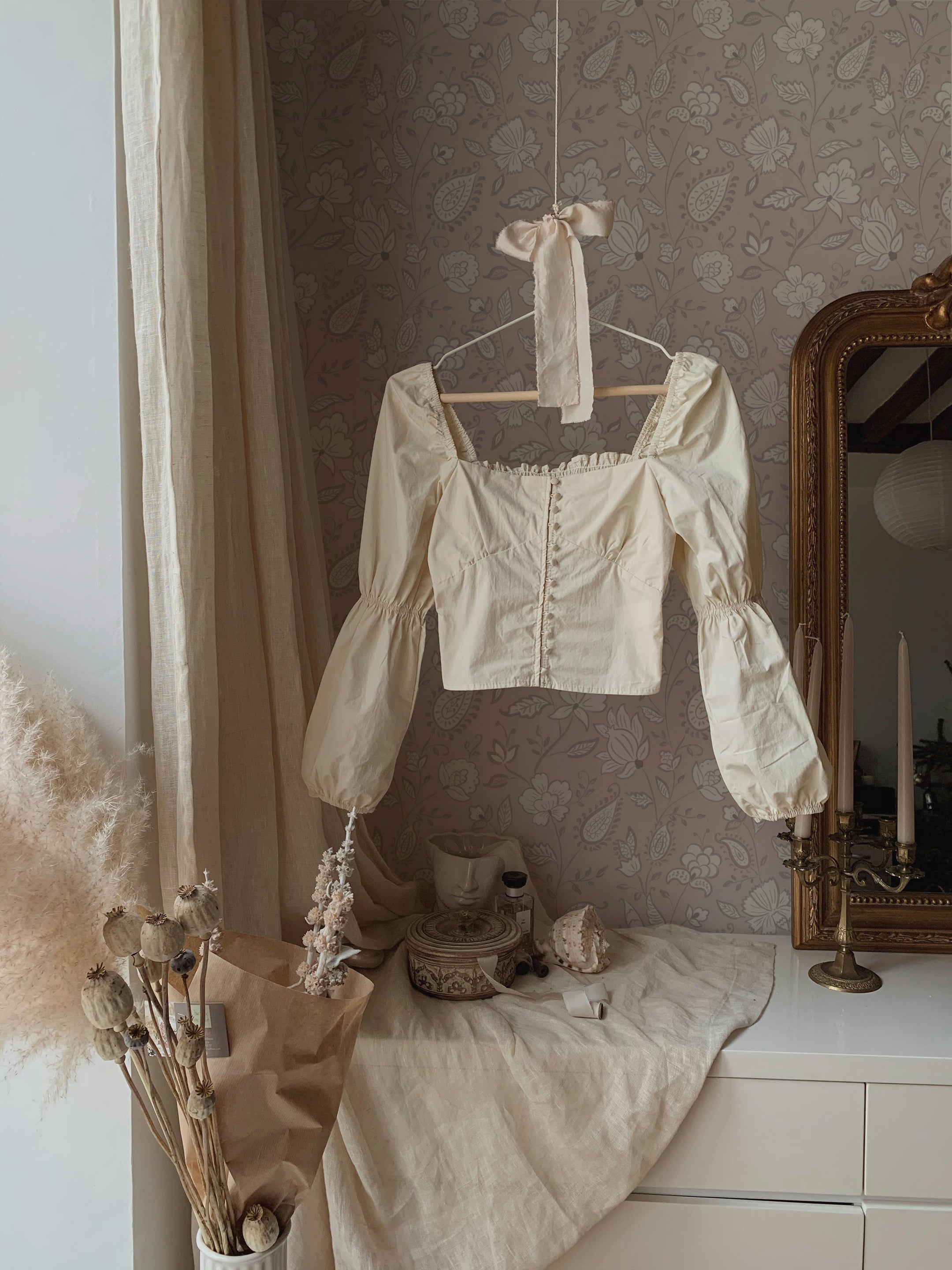 A romantic and rustic styling area featuring a vintage cream blouse hanging on a wooden hanger against a wall covered in soft lavender floral wallpaper. The setting includes dried flowers, decorative objects, and a draped linen fabric, creating an elegant vintage ambiance.