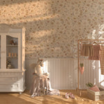 The room features a soft floral wallpaper in pastel tones, a white wardrobe, a unicorn plush toy, and a clothes rack with baby clothes, all bathed in warm sunlight.