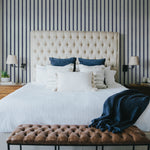 A stylish bedroom with a large tufted beige headboard against a wall covered in navy and white striped wallpaper. The bed is dressed in white linens with navy and white accent pillows and a navy throw blanket. Nightstands with lamps and small decor items flank the bed, creating a cozy and elegant ambiance