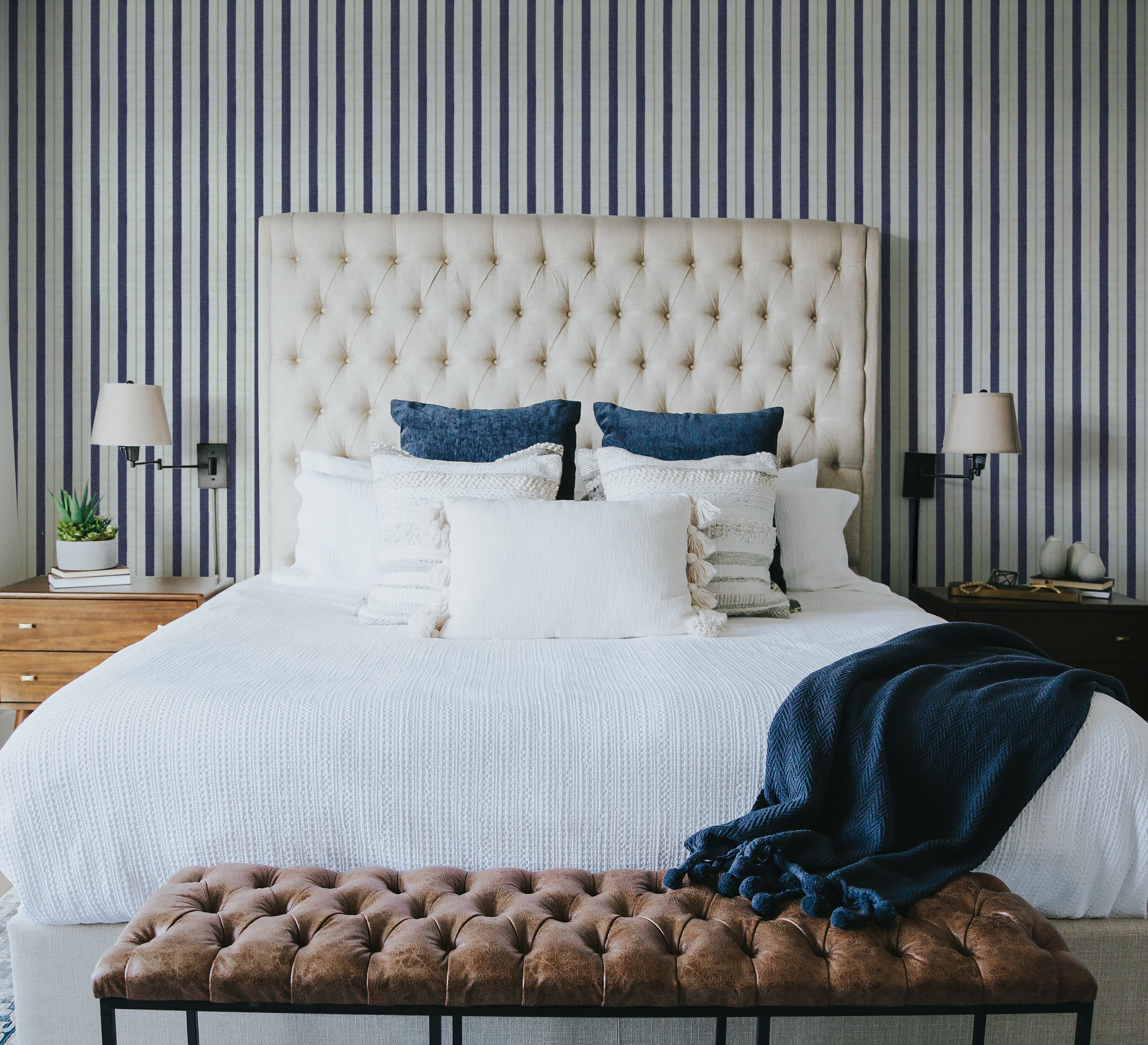 A stylish bedroom with a large tufted beige headboard against a wall covered in navy and white striped wallpaper. The bed is dressed in white linens with navy and white accent pillows and a navy throw blanket. Nightstands with lamps and small decor items flank the bed, creating a cozy and elegant ambiance