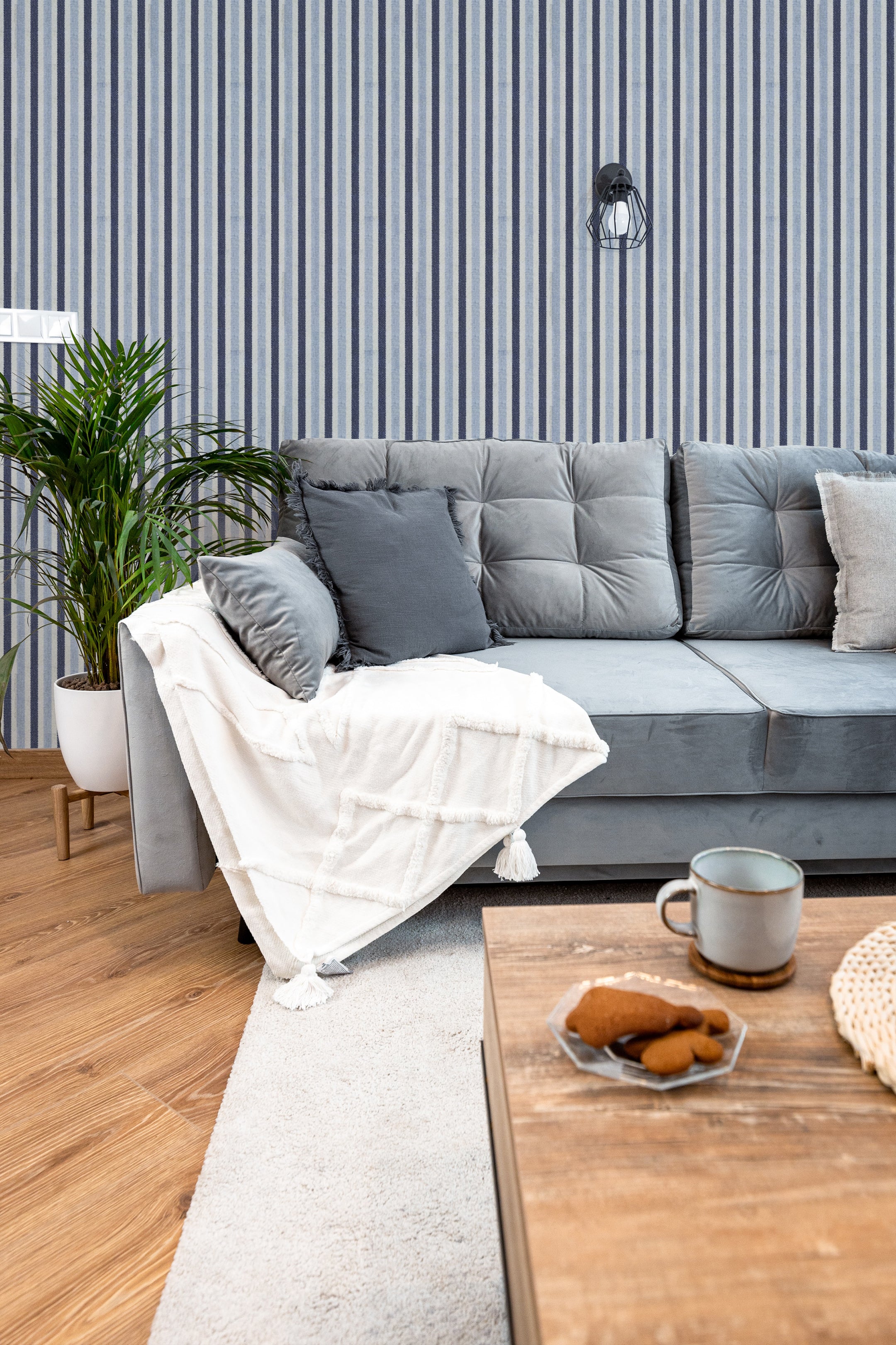 A modern living room with a comfortable gray sofa adorned with matching gray pillows and a white throw blanket. The wall behind the sofa is covered in navy and white striped wallpaper, adding a stylish and contemporary look to the room. A potted plant and a wooden coffee table with a mug and snacks complete the inviting setting.