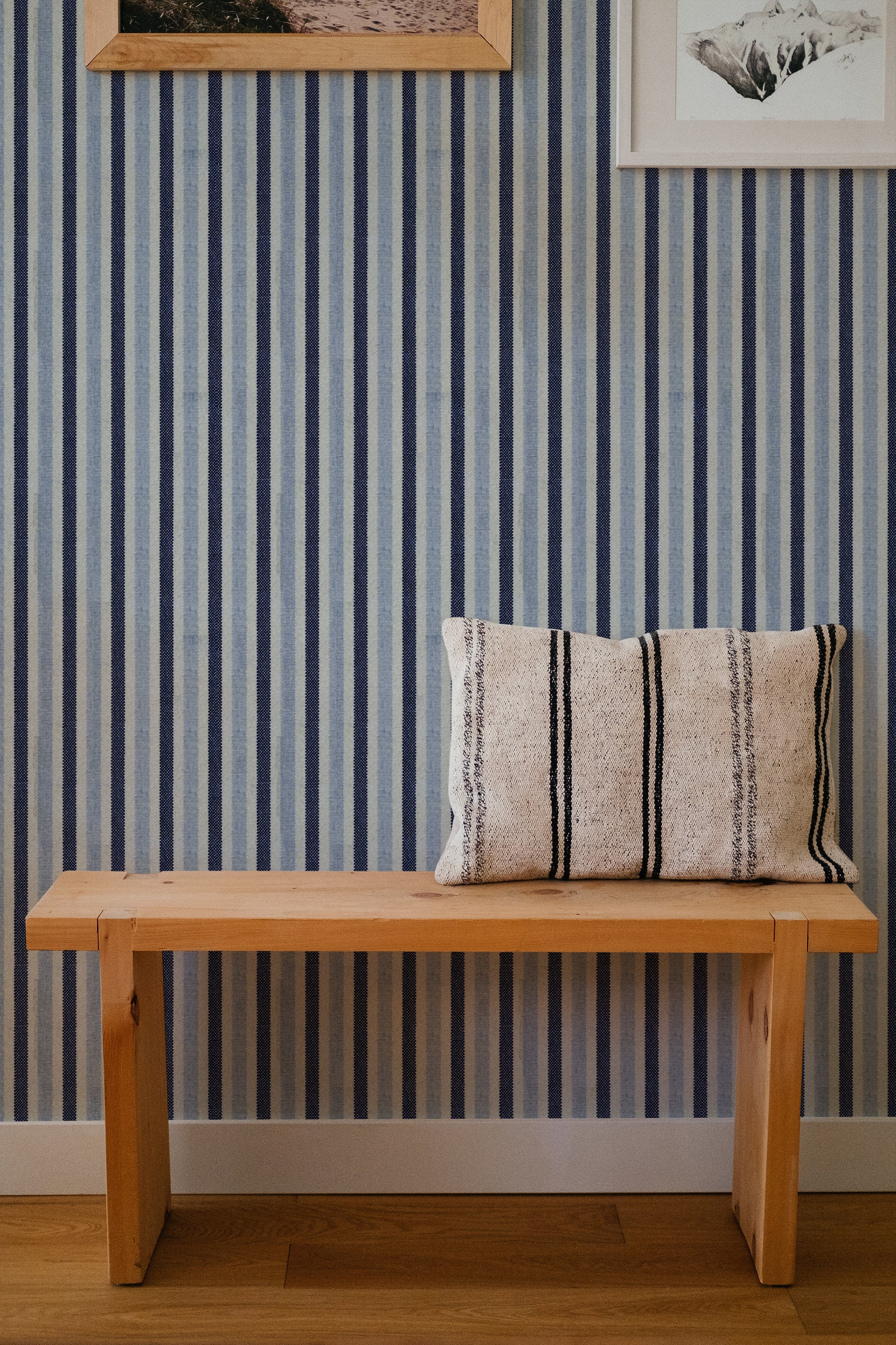 A cozy corner featuring a wooden bench with a striped pillow in front of a wall covered in navy and white striped wallpaper. The wall is decorated with framed pictures, adding a touch of warmth and charm to the space