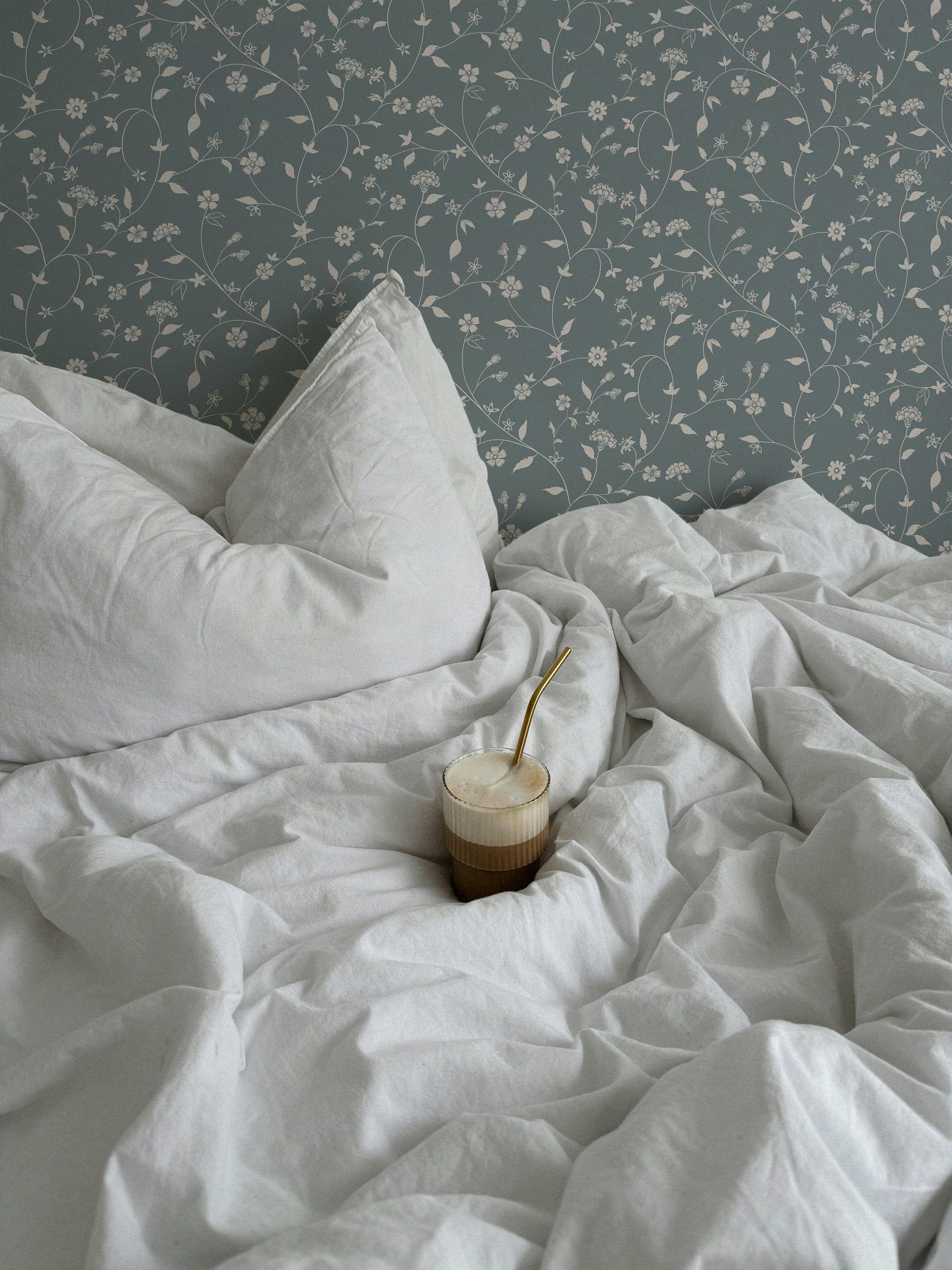 A cozy bedroom scene with white bedding and pillows against a wall covered in Sutton Trailing wallpaper. The wallpaper features a delicate pattern of white floral vines on a blue-gray background. A glass of coffee with a gold straw rests on the rumpled sheets, adding a touch of warmth and relaxation to the serene setting.