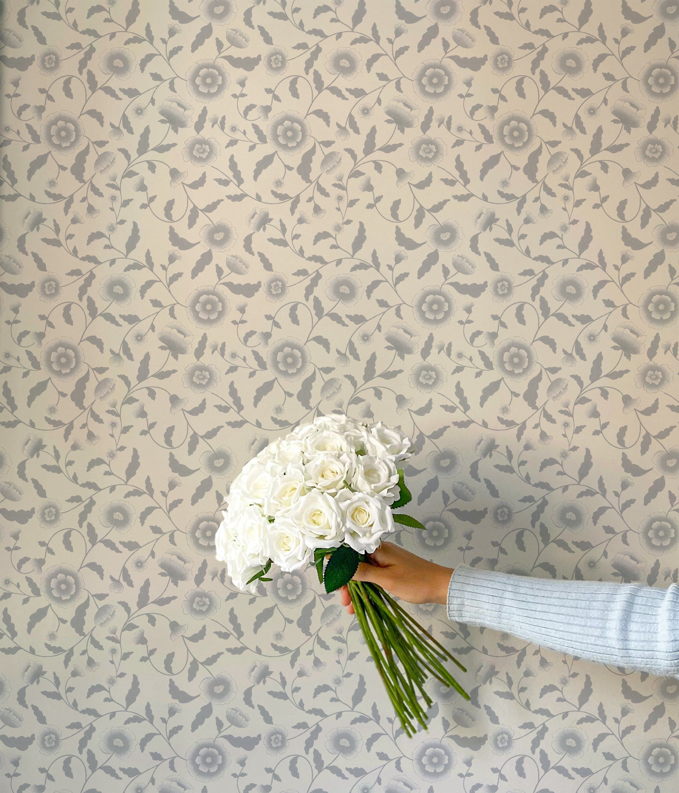 A close-up of a hand holding a bouquet of white roses in front of a wall covered in Frosted Petals wallpaper. The wallpaper displays a pattern of silver floral motifs on a white background, creating a soft and sophisticated backdrop. The combination of the white roses and the delicate wallpaper design adds to the overall sense of elegance and tranquility