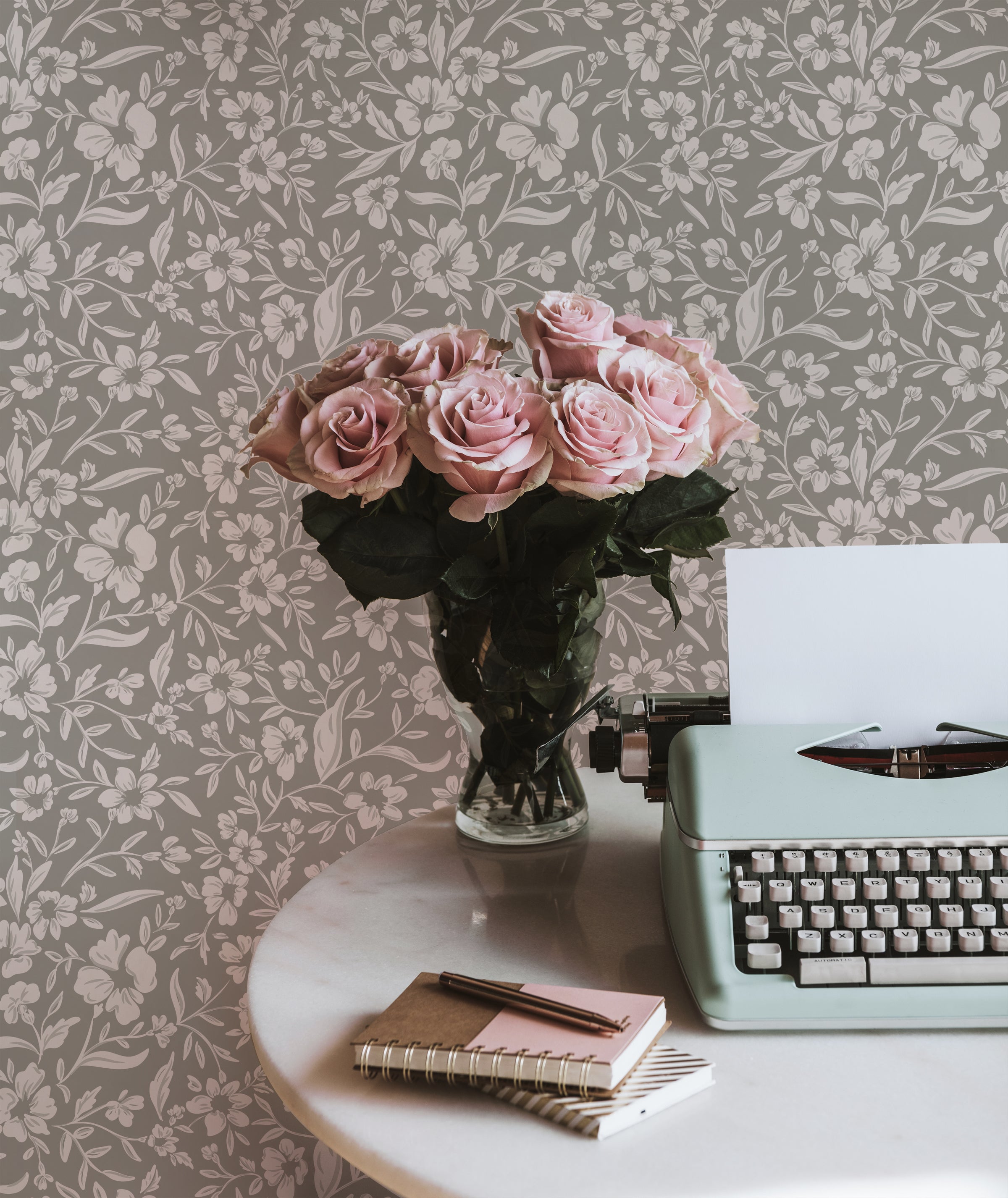 A cozy desk setup with the Soft Meadows Wallpaper in the background, showcasing its elegant floral pattern. A vase of pink roses and a mint green typewriter on a round marble table add a vintage charm.