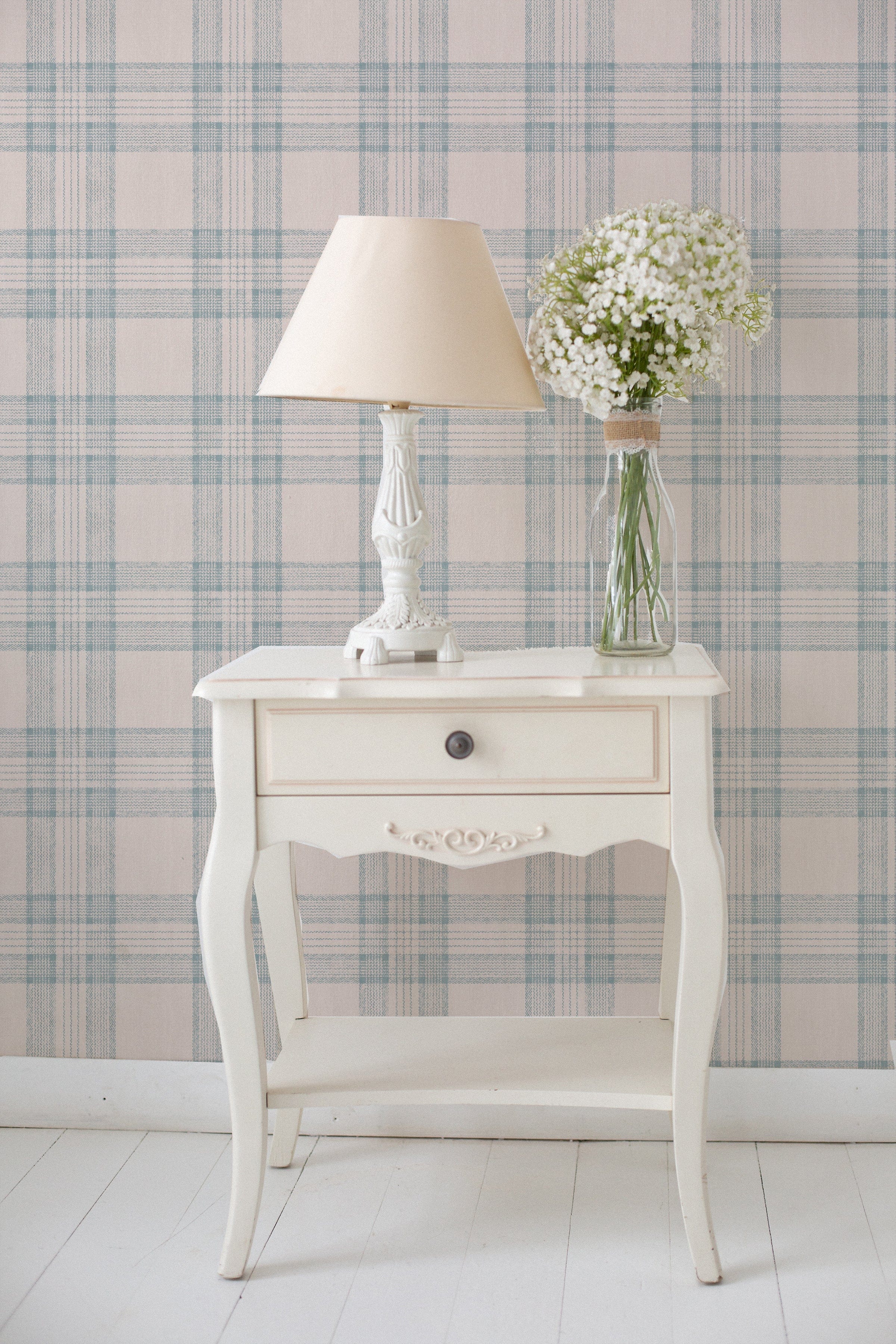 A quaint corner featuring the 'Plaid Wallpaper - Tartan Sand and Sky' as a backdrop, with a classic white side table bearing a traditional lamp with a beige shade and a clear glass vase filled with delicate white baby's breath flowers. The wallpaper's soft plaid pattern complements the vintage charm of the decor, creating a serene and welcoming space.