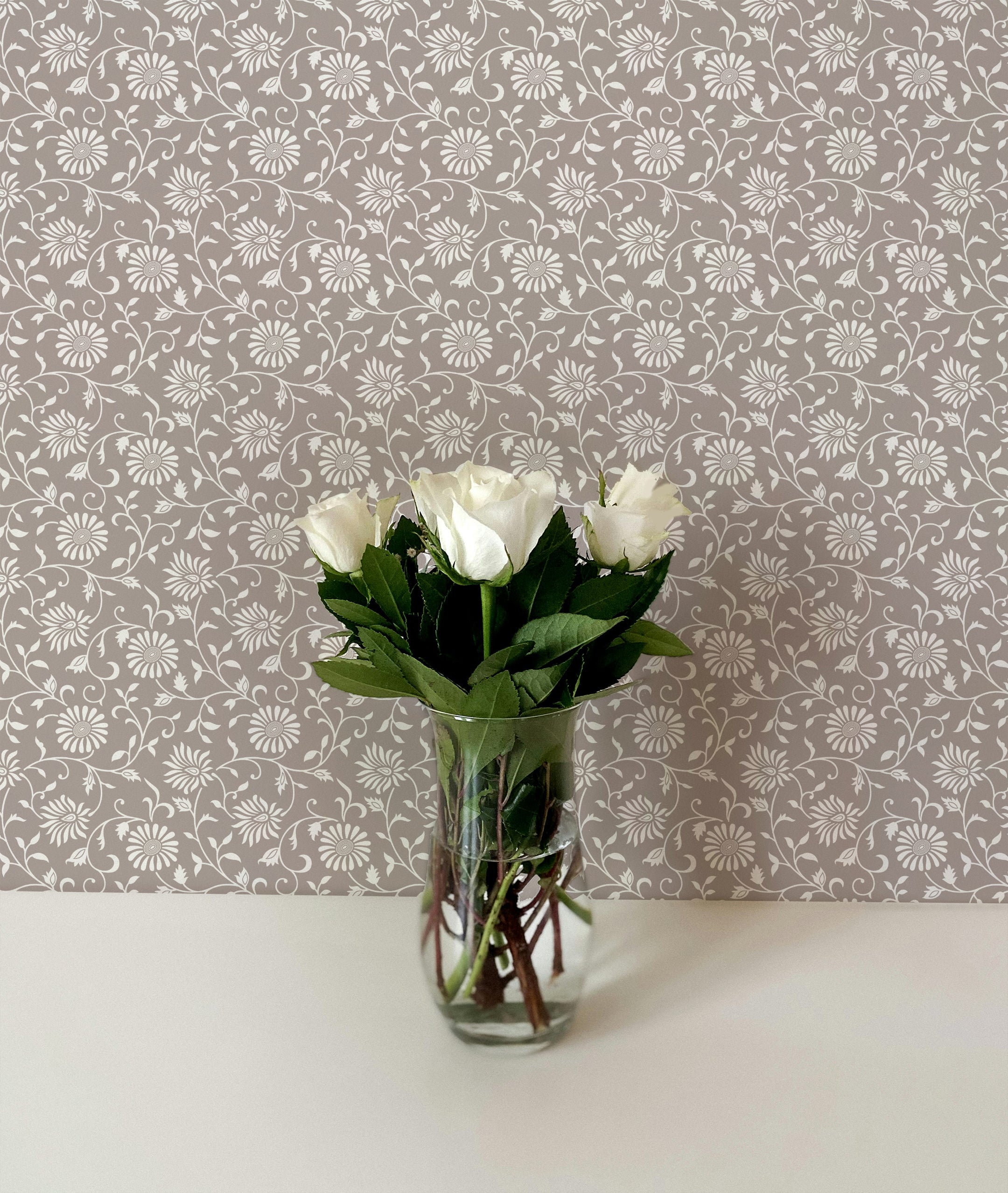 A close-up of a clear glass vase filled with fresh white roses, set against a wall adorned with Estelle Floral Wallpaper. The wallpaper features a pattern of white floral motifs on a soft taupe background, enhancing the timeless and sophisticated look of the space. The combination of the fresh flowers and the vintage wallpaper creates a harmonious and elegant setting.