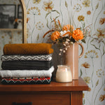A cozy corner of a room showcasing the Warm Glow Floral Wallpaper, adorned with delicate watercolor flowers in warm, earthy tones. The scene includes a stack of folded sweaters and a vintage vase with vibrant orange flowers, enhancing the wallpaper's warm, inviting feel