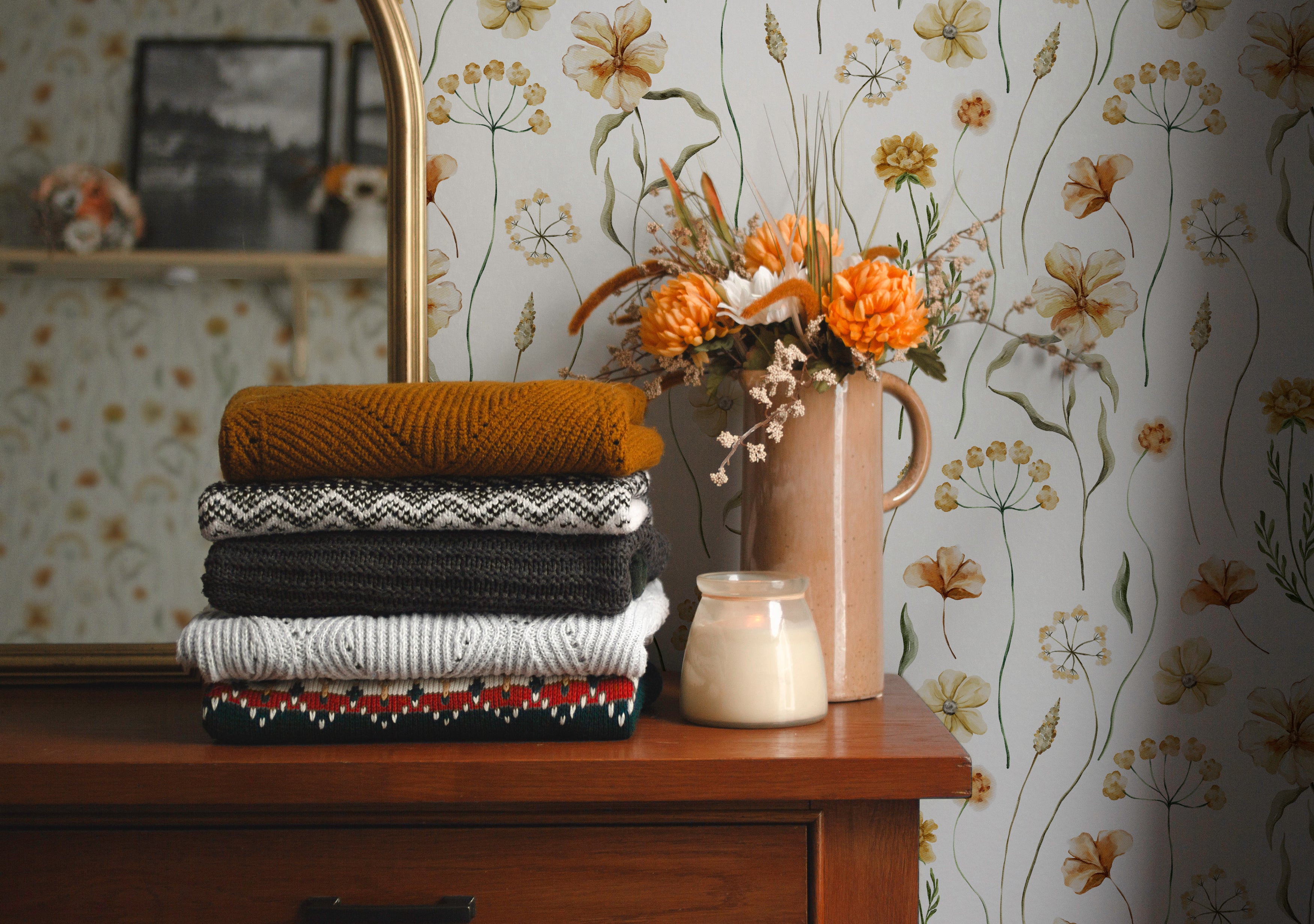A cozy corner of a room showcasing the Warm Glow Floral Wallpaper, adorned with delicate watercolor flowers in warm, earthy tones. The scene includes a stack of folded sweaters and a vintage vase with vibrant orange flowers, enhancing the wallpaper's warm, inviting feel
