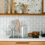 A modern kitchen enhanced by Warm Glow Floral Wallpaper, featuring a subtle and elegant floral pattern. Wooden kitchen accessories and white tiles complement the wallpaper's soft, natural color palette, creating a harmonious and inviting cooking space.