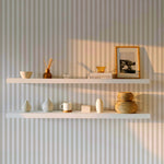 A minimalistic room with Anne Stripe Wallpaper featuring vertical beige and white stripes. Two white floating shelves hold various decor items, including ceramics, books, and a framed photo, with soft sunlight casting shadows.
