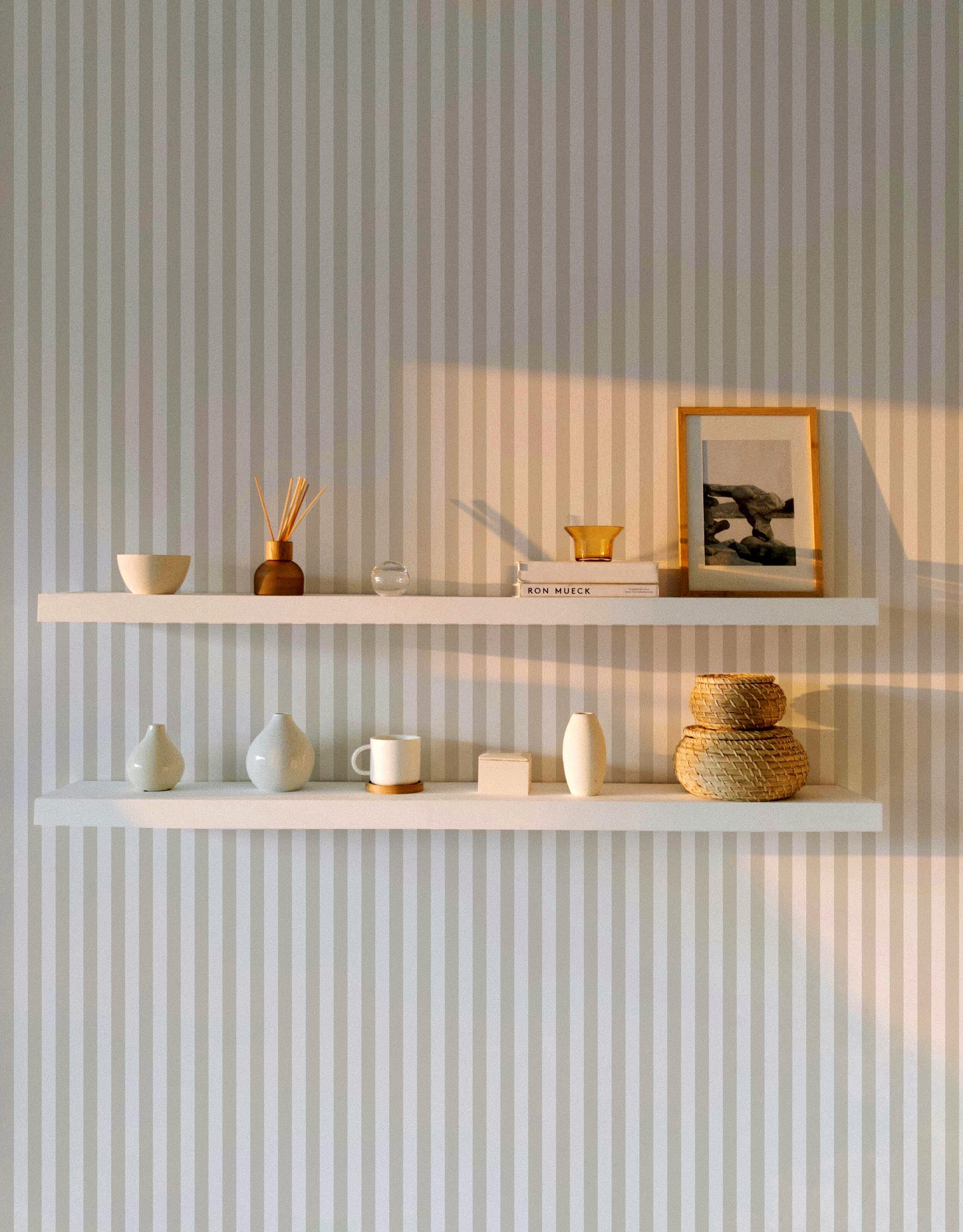 A minimalistic room with Anne Stripe Wallpaper featuring vertical beige and white stripes. Two white floating shelves hold various decor items, including ceramics, books, and a framed photo, with soft sunlight casting shadows.