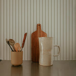 A kitchen counter scene with Anne Stripe Wallpaper in the background. The counter has a ceramic mug, a wooden cutting board, and a container holding wooden utensils, enhancing the warm and cozy atmosphere.