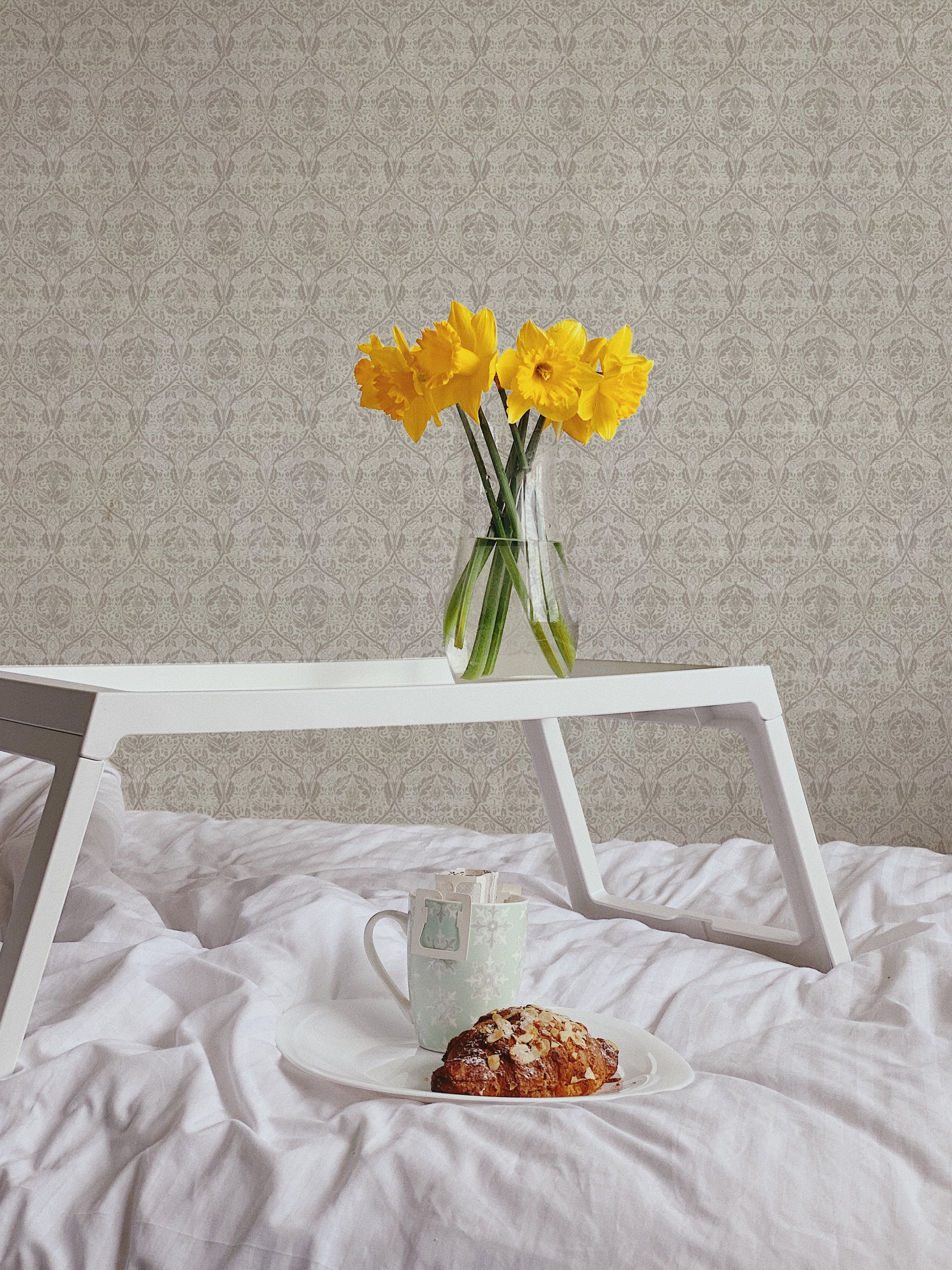 A cozy room setup featuring a white side table with a clear vase holding vibrant yellow daffodils. A white mug with a floral design and a scone on a plate are placed next to the vase, all atop a textured white bedspread. The room is decorated with an elegant gray and beige floral patterned wallpaper, providing a subtle and classic backdrop.