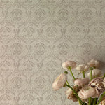A delicate floral wallpaper in gray and beige hues, displayed behind a vase of light pink ranunculus flowers, partially wilted, adding a touch of natural beauty to the scene. The wallpaper's classic design features detailed flowers and leaves, contributing to a serene and refined ambiance.