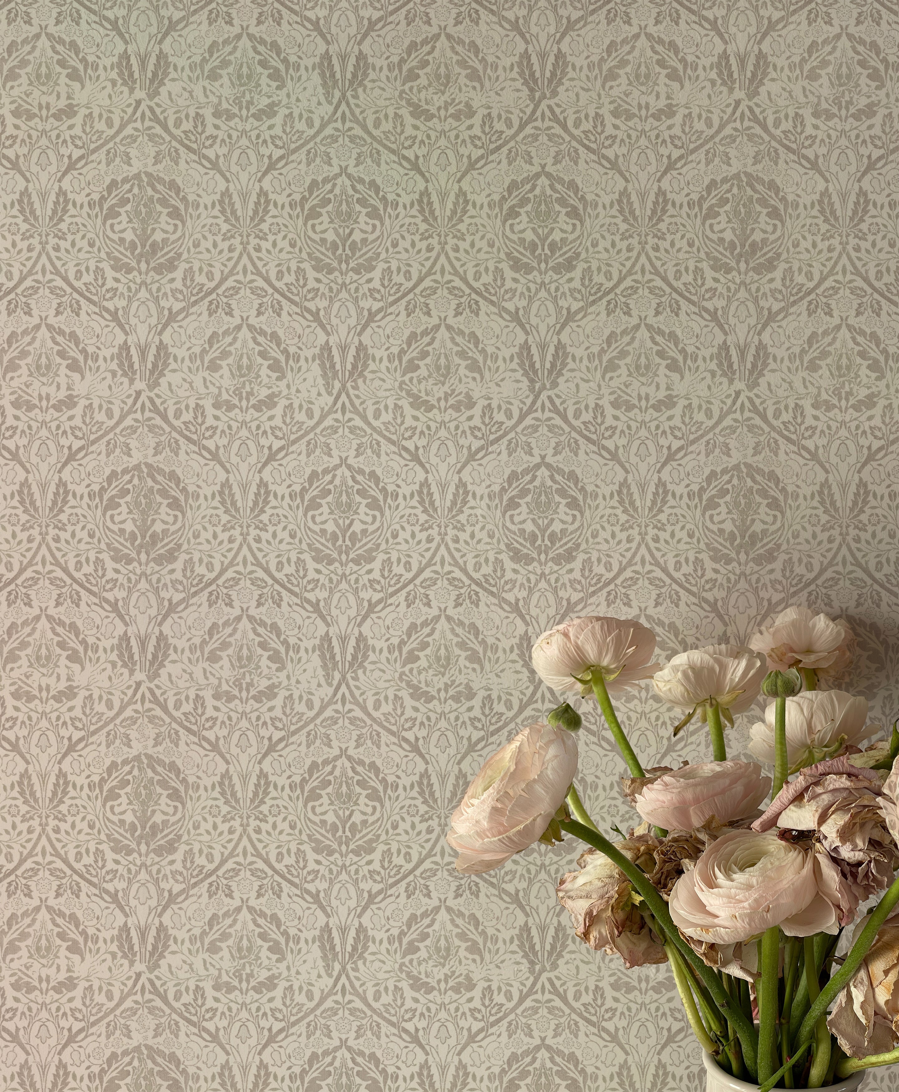 A delicate floral wallpaper in gray and beige hues, displayed behind a vase of light pink ranunculus flowers, partially wilted, adding a touch of natural beauty to the scene. The wallpaper's classic design features detailed flowers and leaves, contributing to a serene and refined ambiance.