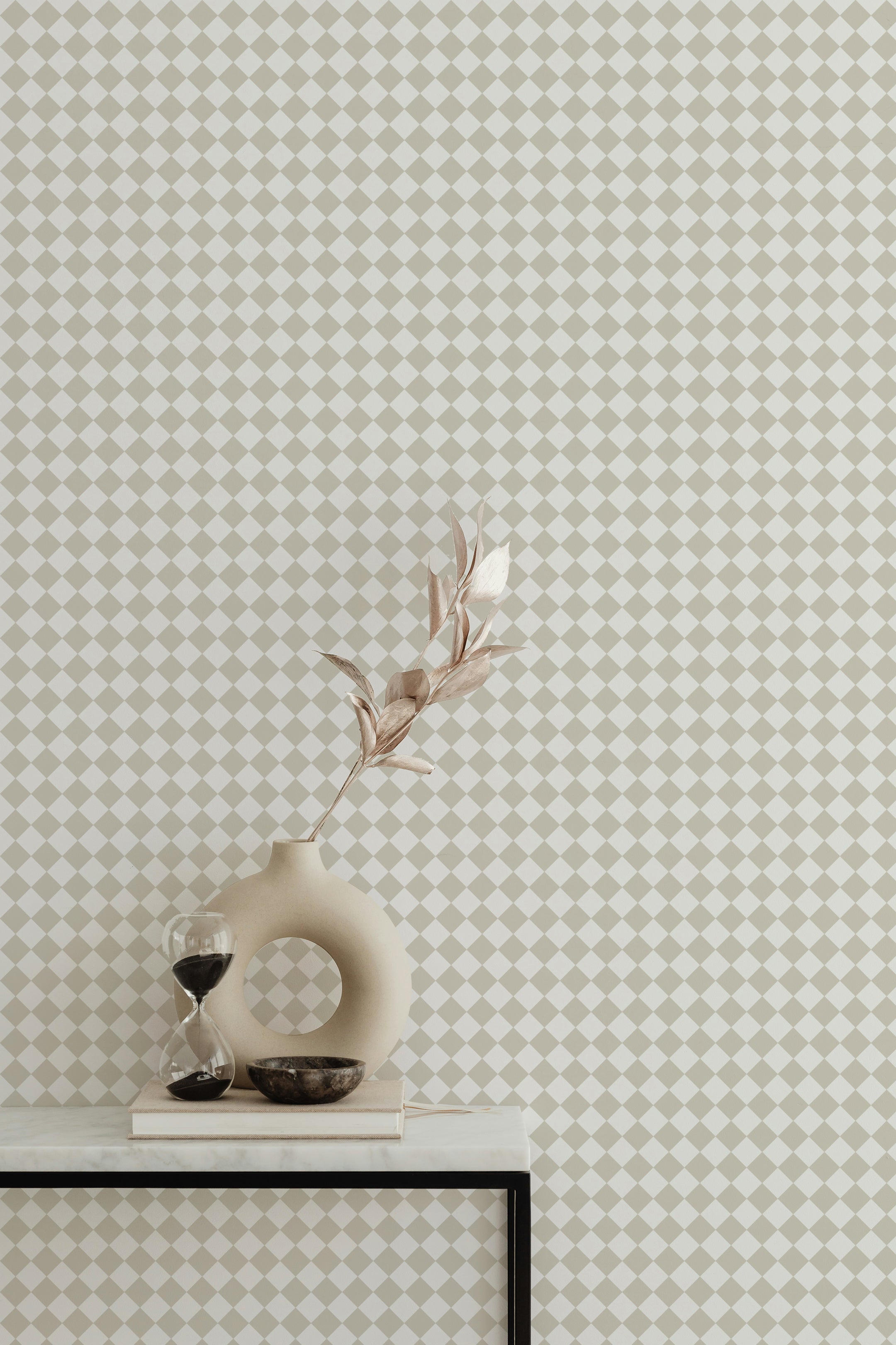 A stylish decor arrangement with a beige sculptural vase holding a dried leaf stem, an hourglass, and a small bowl placed on a white surface. The wall behind features beige and white checkered wallpaper, enhancing the space with its classic and refined pattern