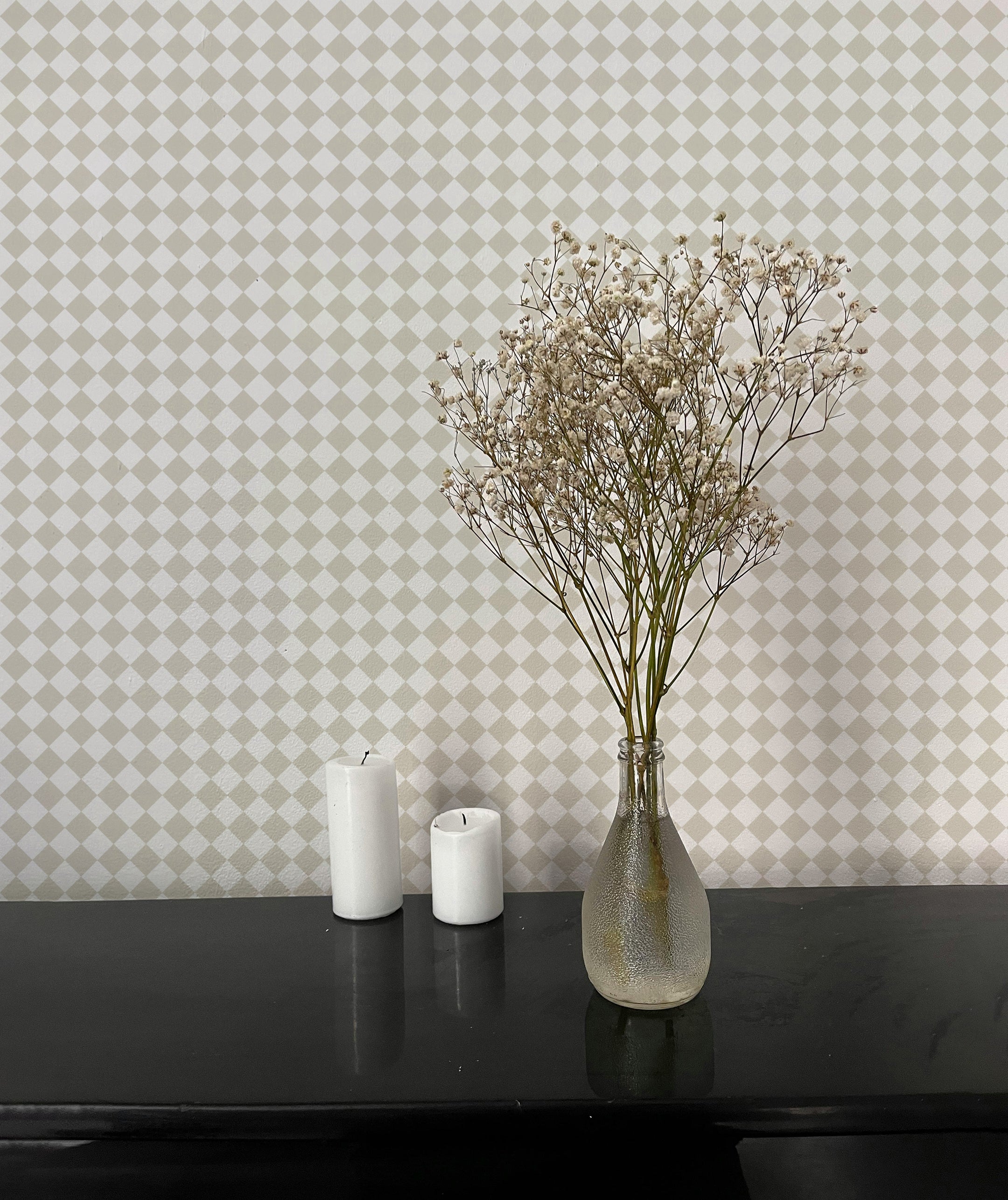 A minimalist setting featuring a black surface with two white candles and a clear vase holding delicate dried flowers. The background showcases beige and white checkered wallpaper, adding a subtle and elegant pattern to the scene.