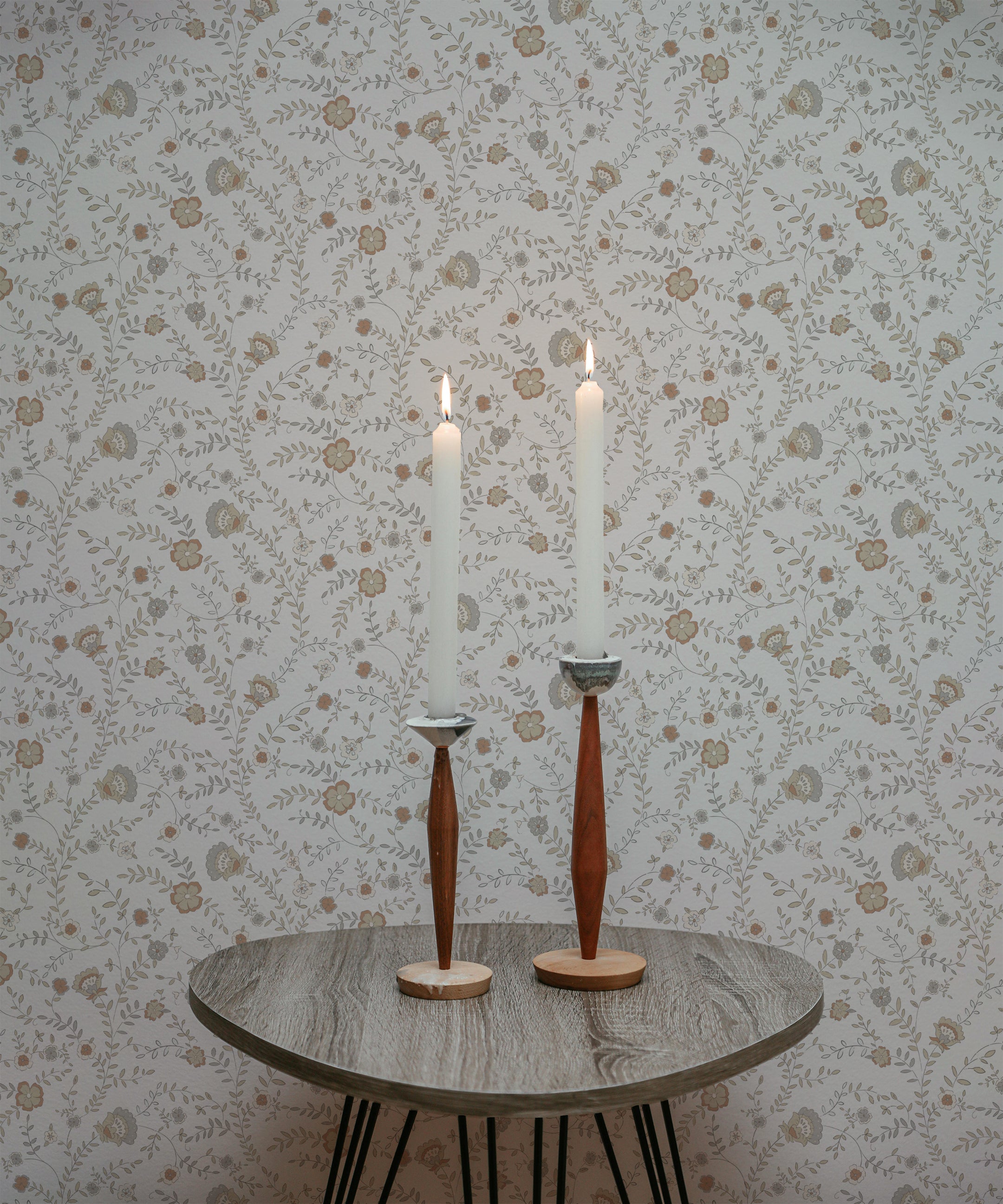 A close-up view of a small round wooden table with hairpin legs, positioned against a wall covered in Boho Trailing Foliage wallpaper. The table holds two lit white taper candles in tall, slender wooden candle holders. The wallpaper features a delicate pattern of small, pastel-colored flowers and leaves, adding a soft, natural ambiance to the setting