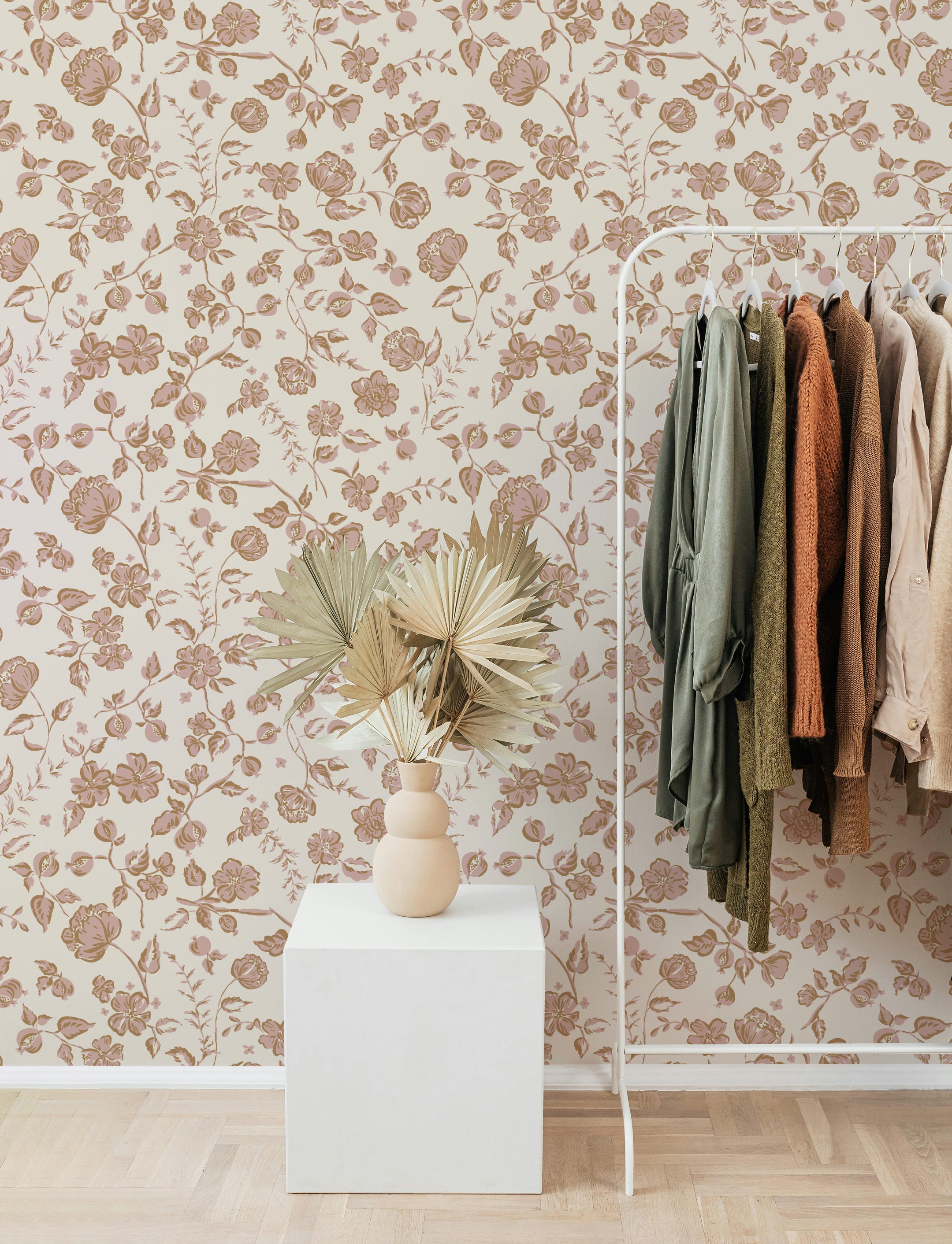 A stylish room with the Rosy Elegance Wallpaper covering the wall. A white clothing rack with various earthy-toned garments stands beside a white cube table with decorative dried palm leaves in a beige vase