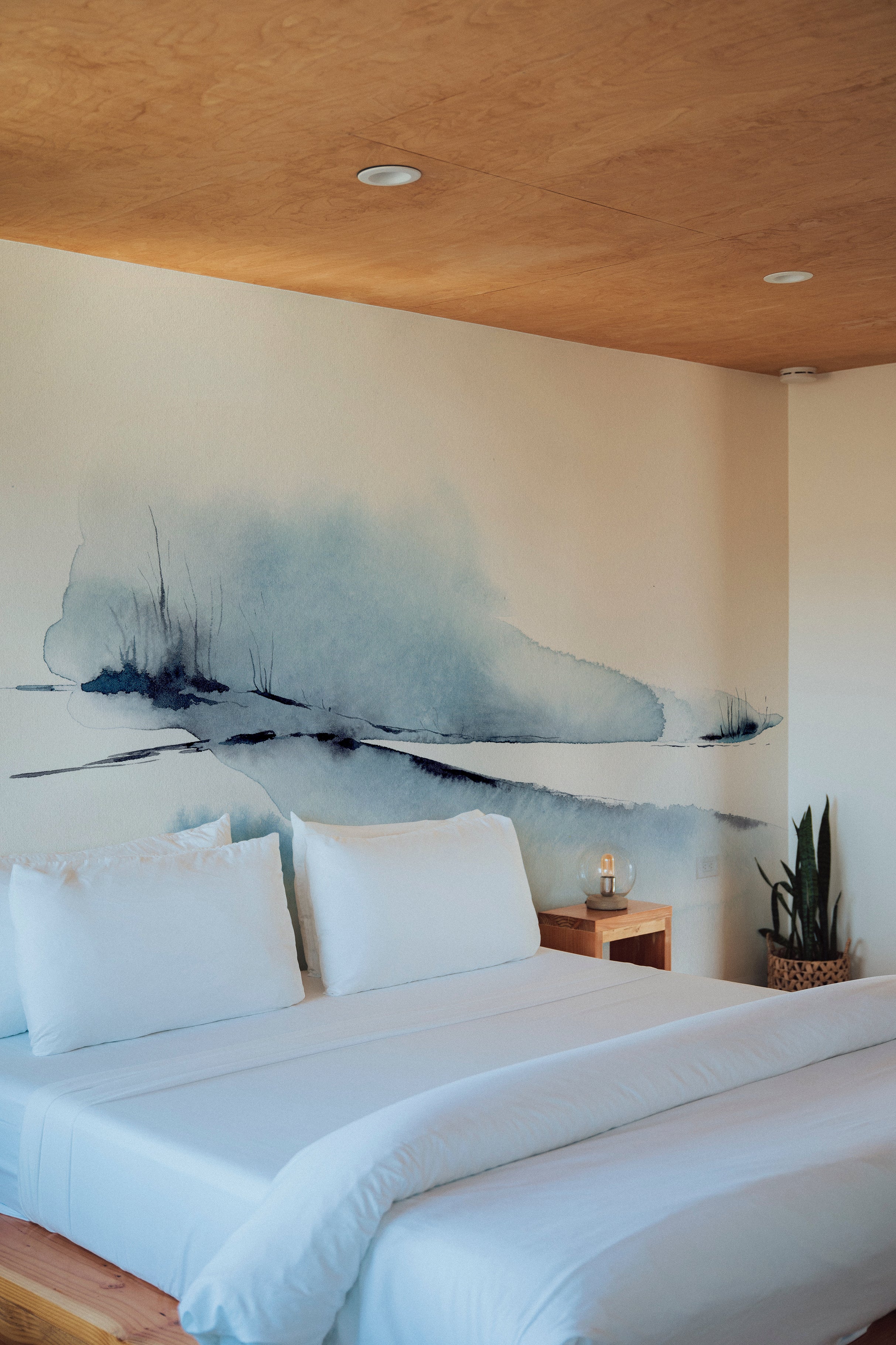 A serene bedroom setup with the "Atmospheric Sky" wall mural painting an ethereal watercolor landscape behind the bed. The mural's soothing blues and detailed brush strokes create a peaceful ambiance, complemented by simple white bedding and minimalistic decor.