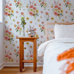 Golden Garden Wallpaper with a vibrant floral pattern featuring pink, yellow, and white flowers, enhancing the wall behind a wooden bedside table with a clock and a small flower vase in a bright bedroom