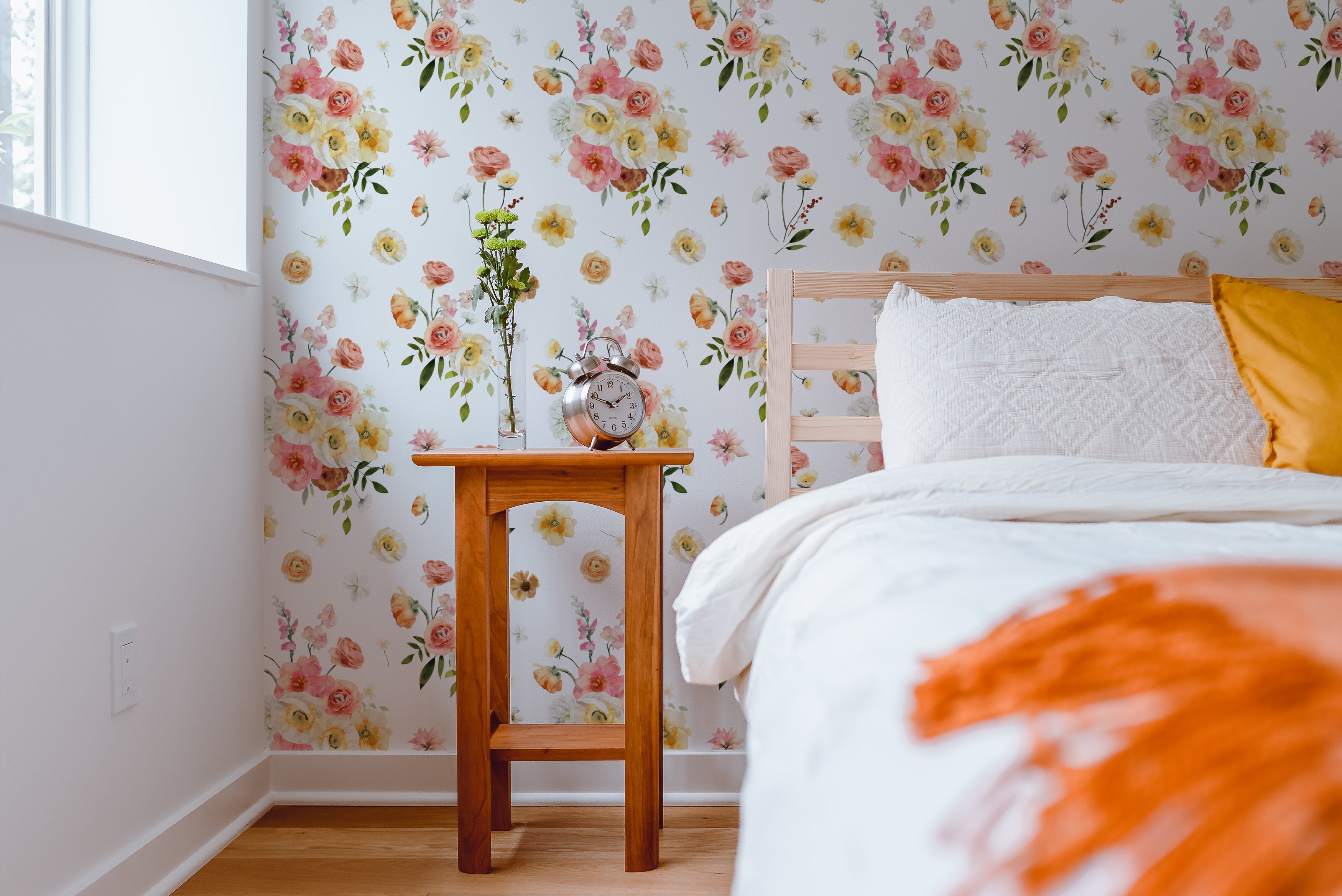 Golden Garden Wallpaper with a vibrant floral pattern featuring pink, yellow, and white flowers, enhancing the wall behind a wooden bedside table with a clock and a small flower vase in a bright bedroom