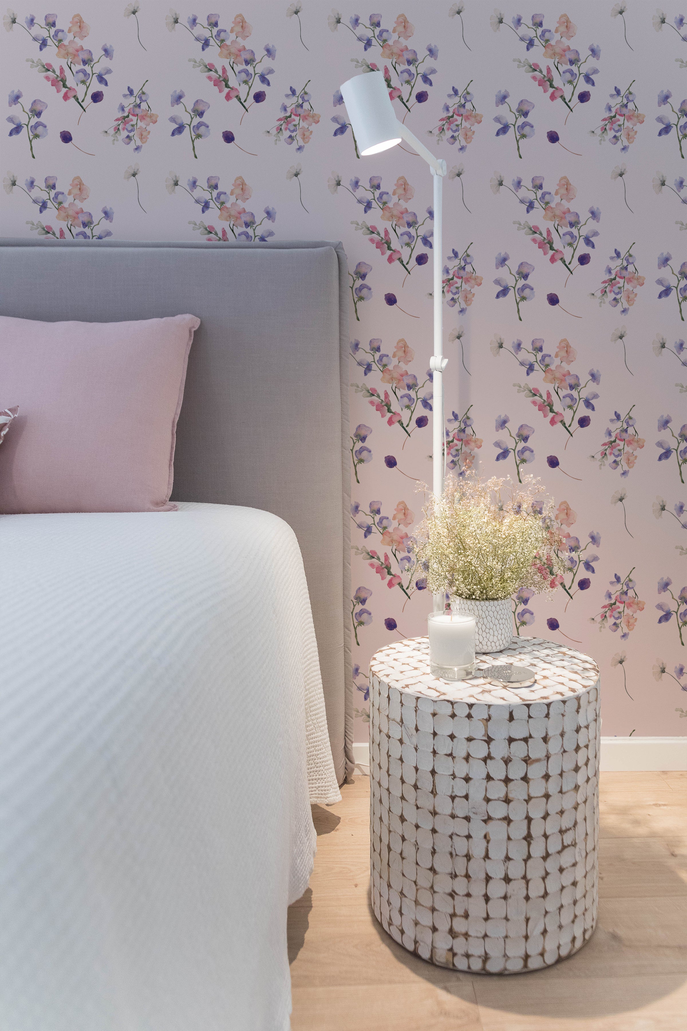 Violet Bloom Wallpaper featuring a soft floral pattern on a nude background in a bedroom setting, with a grey headboard, pink pillows, and a white bedside lamp and table.