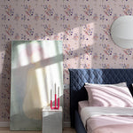 Violet Bloom Wallpaper in a bedroom setting with a blue quilted headboard, pink bedding, and a painting leaning against the wall, highlighting the floral design on a nude background.