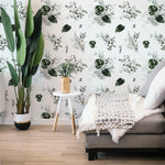 A well-appointed living space showcasing the Tropical Greenery Wallpaper, featuring an array of lush green botanical prints including monstera leaves and fine herbs against a soft white background. A cozy beige sofa adorned with a textured throw and complemented by a large indoor plant enhances the room's natural vibe.