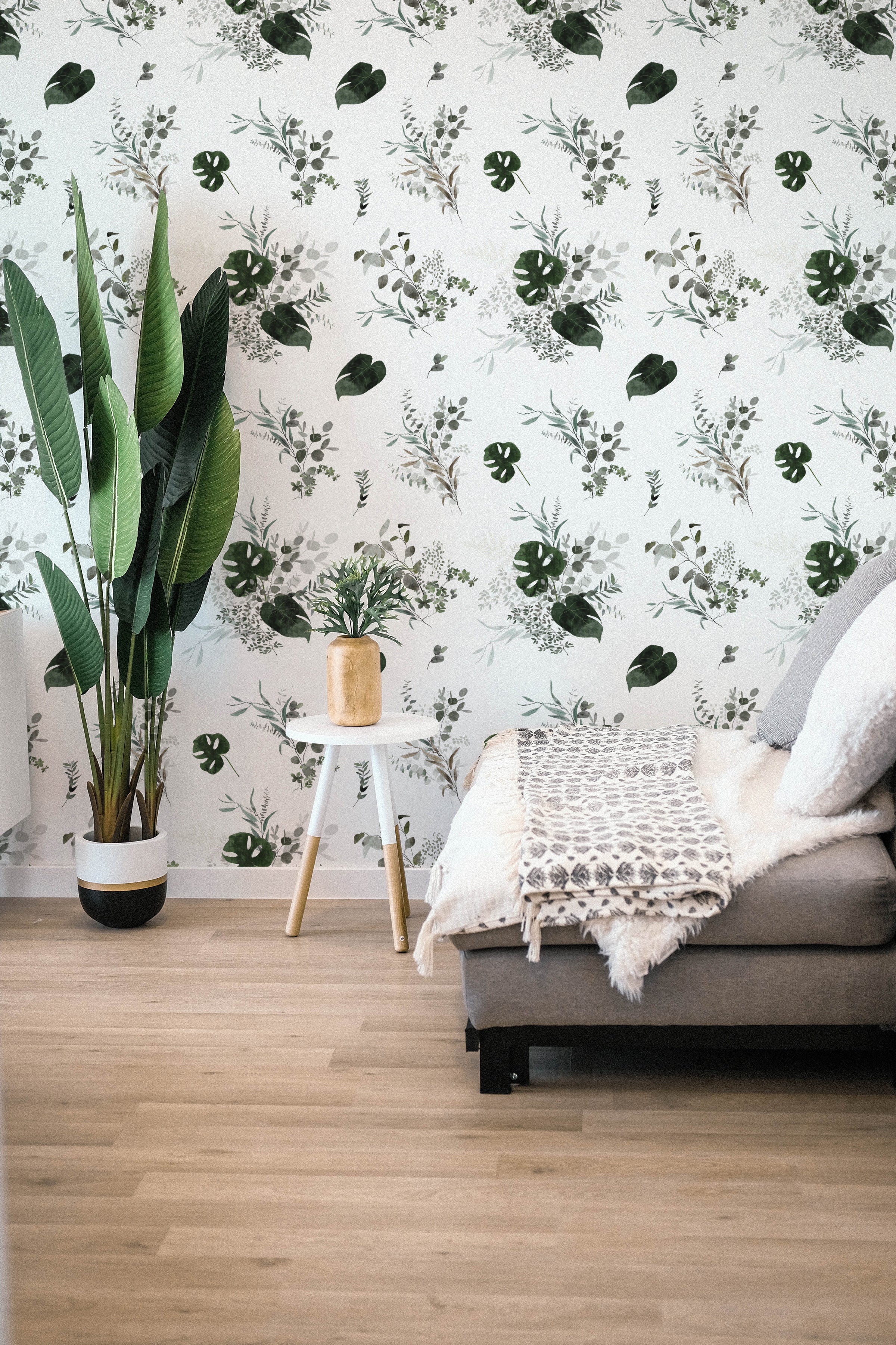 A well-appointed living space showcasing the Tropical Greenery Wallpaper, featuring an array of lush green botanical prints including monstera leaves and fine herbs against a soft white background. A cozy beige sofa adorned with a textured throw and complemented by a large indoor plant enhances the room's natural vibe.