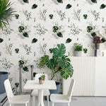 A vibrant dining area with the Tropical Greenery Wallpaper providing a lively backdrop. The space features white furniture, including a simple table and chairs, accented with indoor plants that echo the wallpaper's tropical theme, creating a harmonious indoor-outdoor feel.