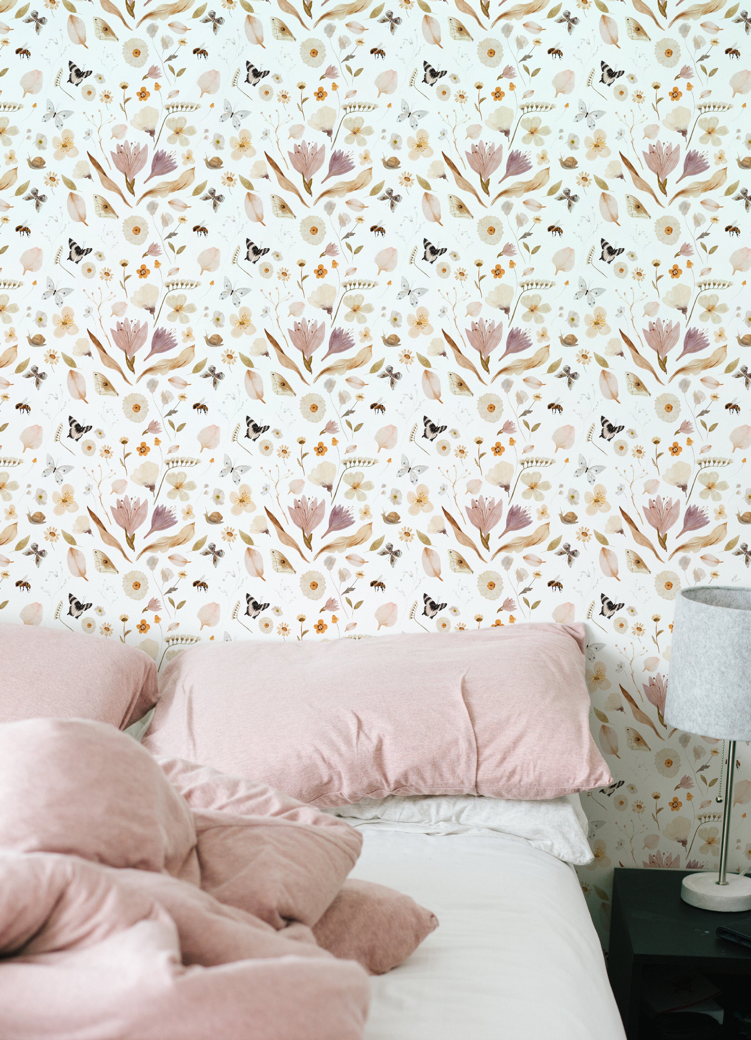 A square image displaying the Boho Garden Wallpaper in a bedroom setting. The floral pattern creates a focal point behind a bed with blush-colored pillows, complementing the relaxed bohemian aesthetic of the room.