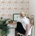 A vertical image capturing a cozy home office with the Boho Garden Wallpaper adorning the wall. The wallpaper features a vibrant pattern of watercolor wildflowers, leaves, and butterflies in shades of pink, gold, and green. A person with their back to the camera sits at a white desk, contemplating the wallpaper, amidst a peaceful setting with indoor plants and soft, natural lighting.