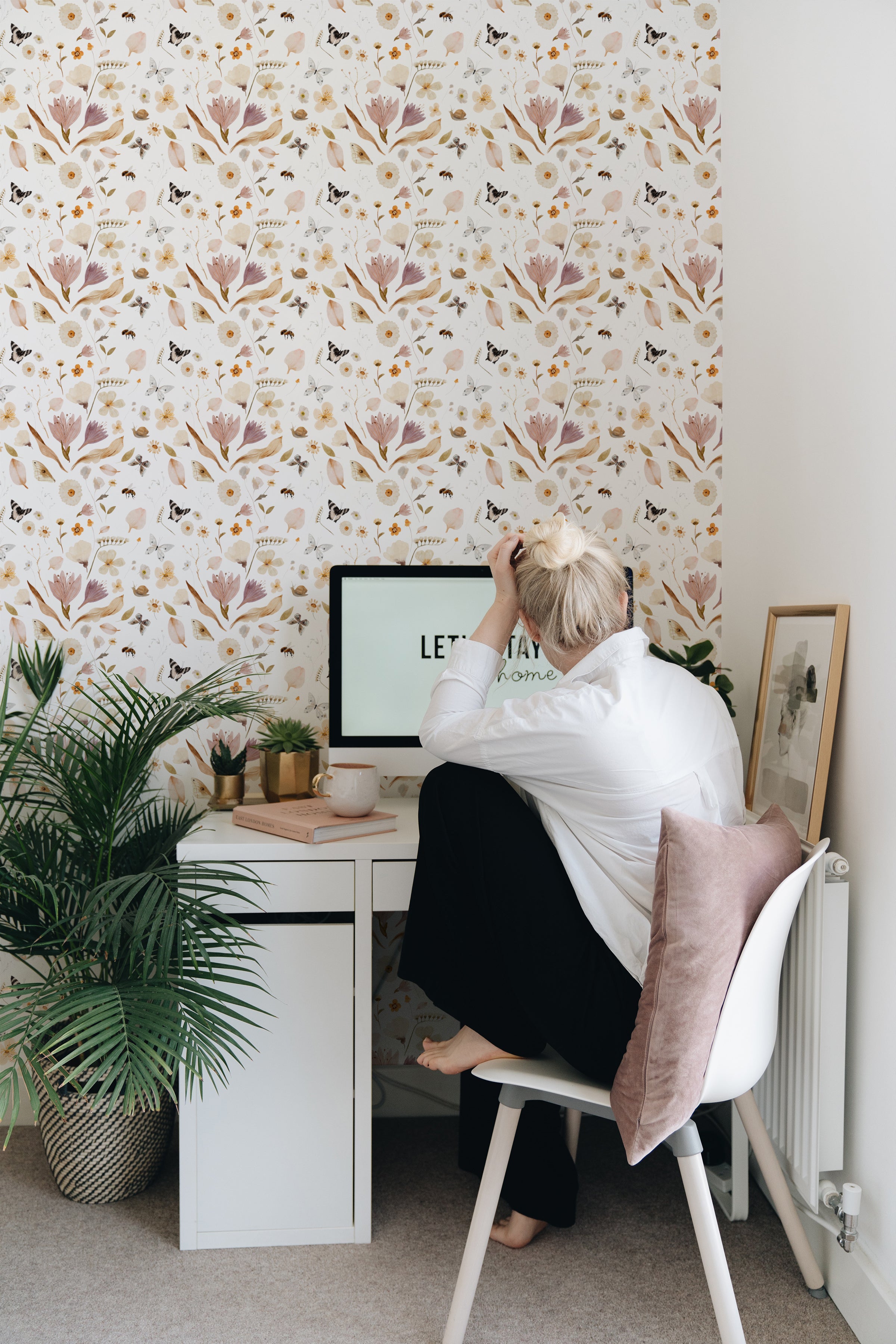 A vertical image capturing a cozy home office with the Boho Garden Wallpaper adorning the wall. The wallpaper features a vibrant pattern of watercolor wildflowers, leaves, and butterflies in shades of pink, gold, and green. A person with their back to the camera sits at a white desk, contemplating the wallpaper, amidst a peaceful setting with indoor plants and soft, natural lighting.